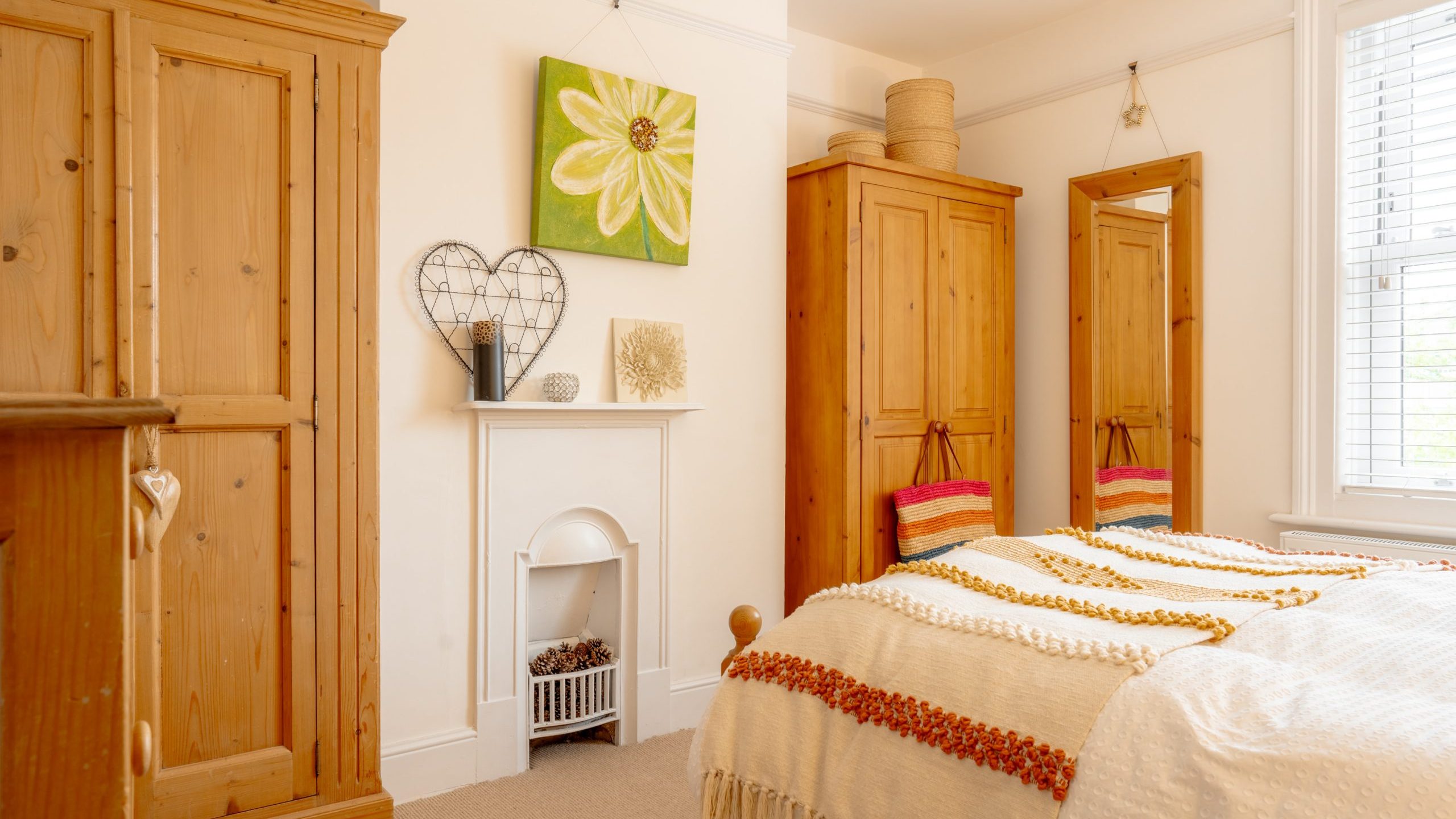A cozy bedroom featuring a bed with a colorful striped blanket, wooden wardrobes, decorative baskets, and a floral artwork above a white fireplace.