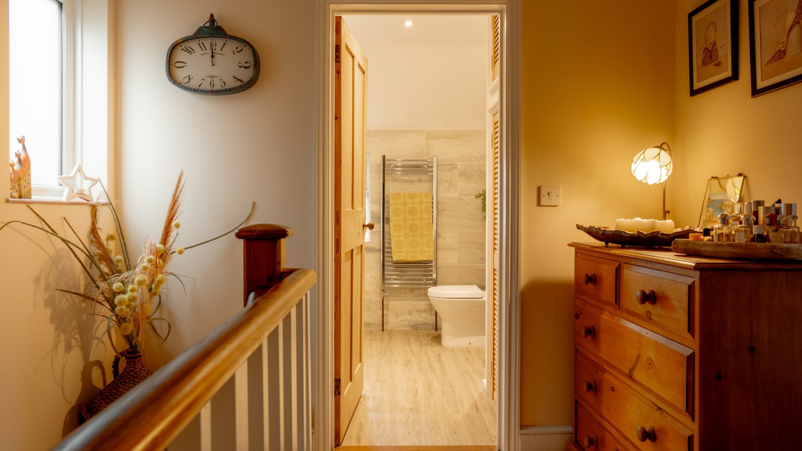 A warmly lit hallway in a home with wooden furniture, pictures on the walls, and an open door leading to a bathroom with visible toilet and shower.