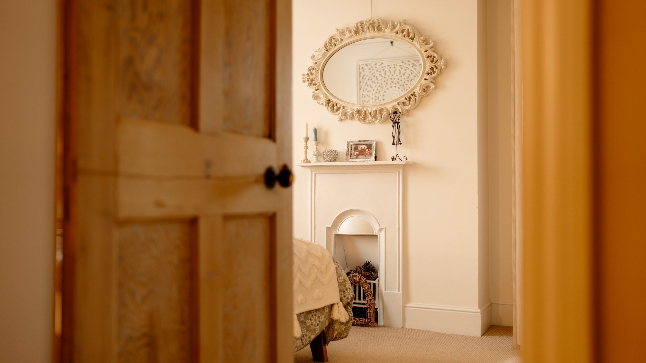 A warmly lit bedroom viewed through a partially open door, featuring an ornate mirror above a fireplace with decorative items on the mantelpiece.