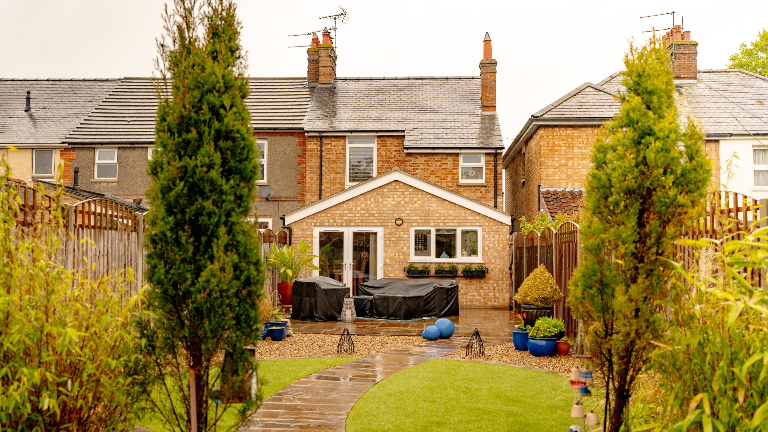A cozy suburban home with an extension and a green lawn, surrounded by fences and lush plants, showcasing a stone path leading to the house under a cloudy sky.