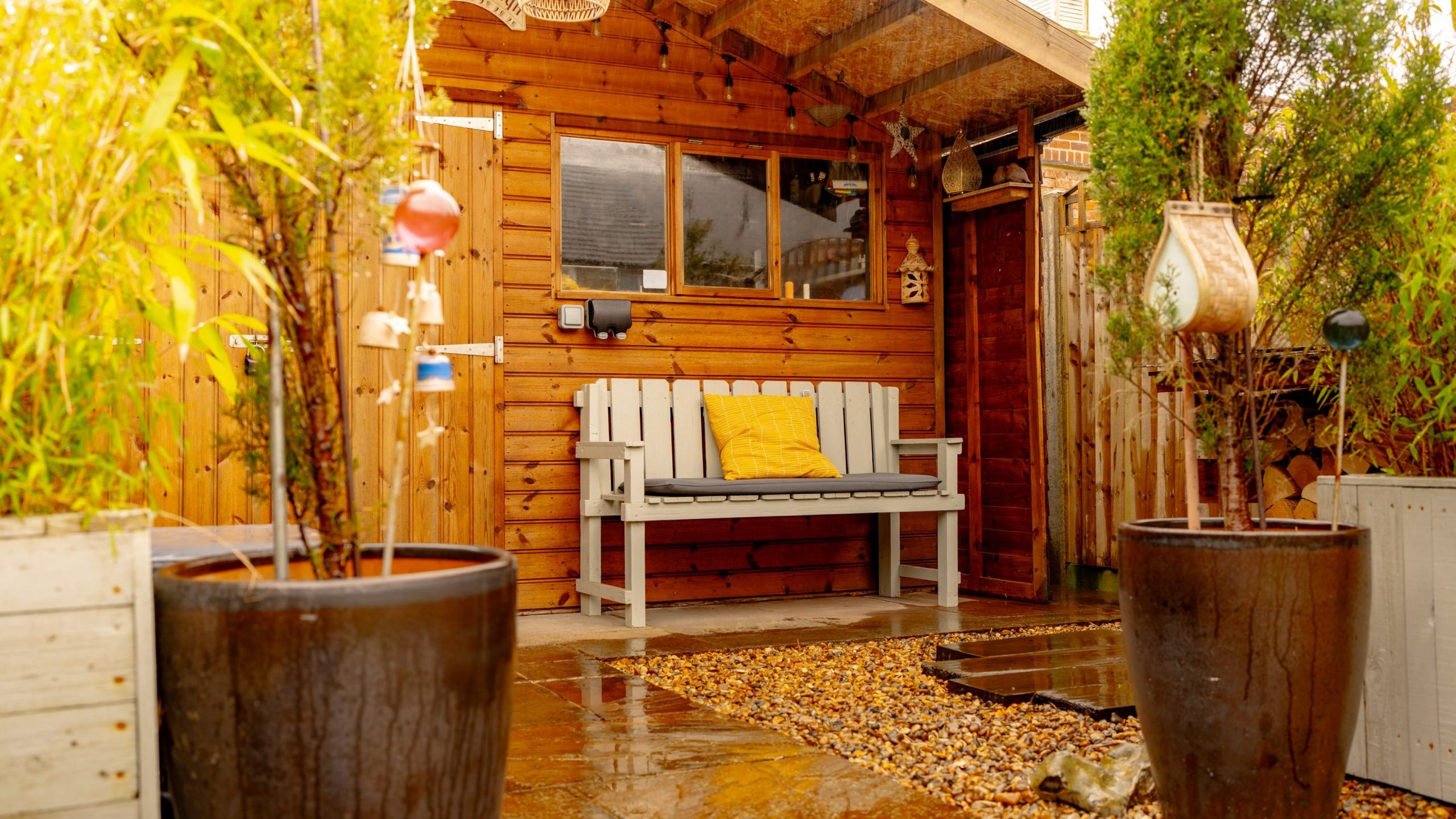 A cozy wooden porch with a bench adorned with yellow cushions, surrounded by green plants and large pots under a gentle rainfall, exuding a warm, inviting ambiance.
