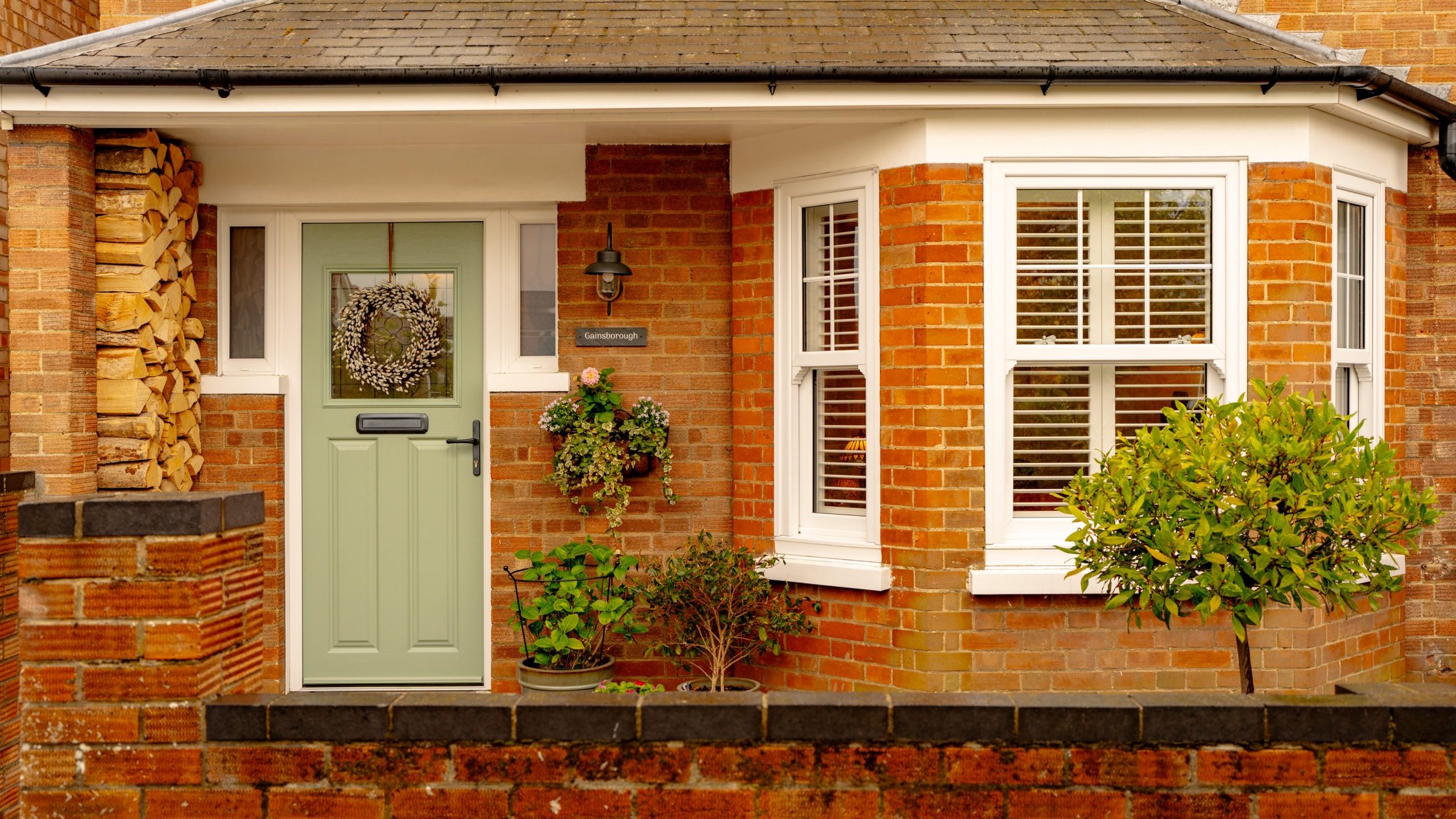 A charming brick house facade with a sage green door, two windows, hanging plants, and neatly stacked firewood alongside a decorative wreath on the door.