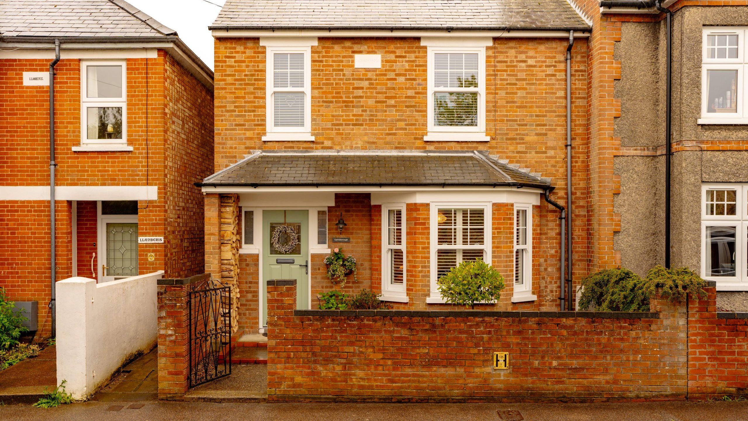 A two-story brick house with white windows and doors, featuring a small front garden enclosed by a low brick wall and a metal gate, on a residential street.
