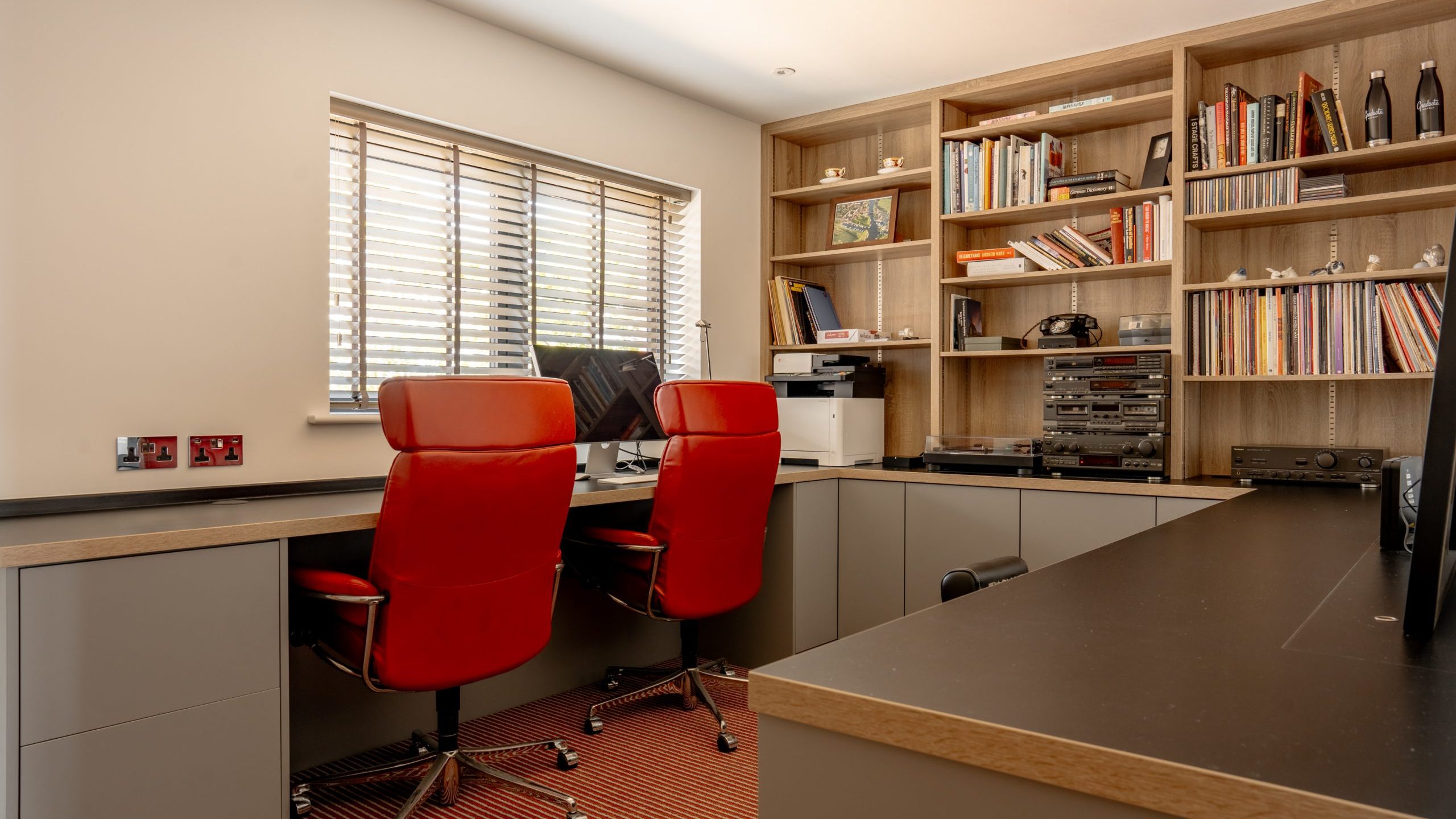 A modern home office with a large wooden desk, two red leather office chairs, and a shelving unit filled with books, vinyl records, and various decor items. The room is well-lit by natural light coming through a window with horizontal blinds.