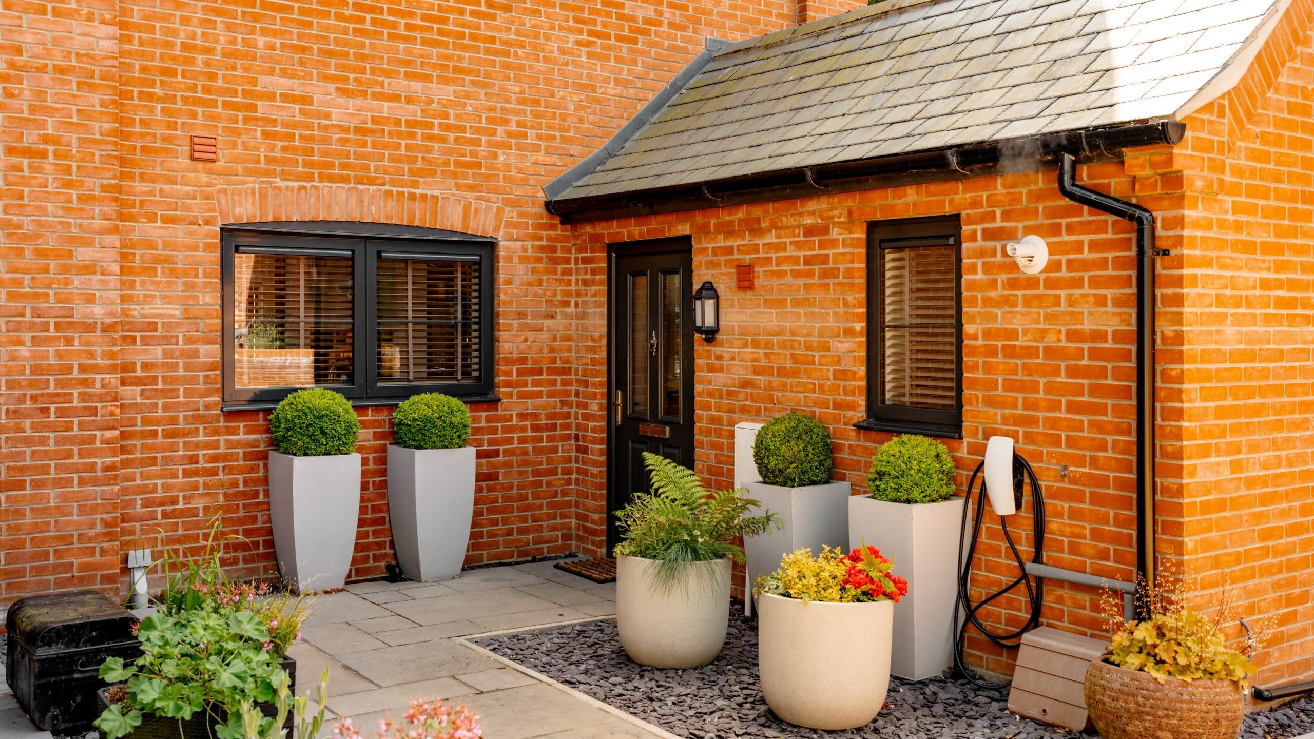 A neat courtyard with brick walls features modern black-framed windows, a black door, and a small roofed entrance. Potted plants, including topiaries and flowers, adorn the space. A white wall light and a coiled hose are attached to the wall near the entrance.