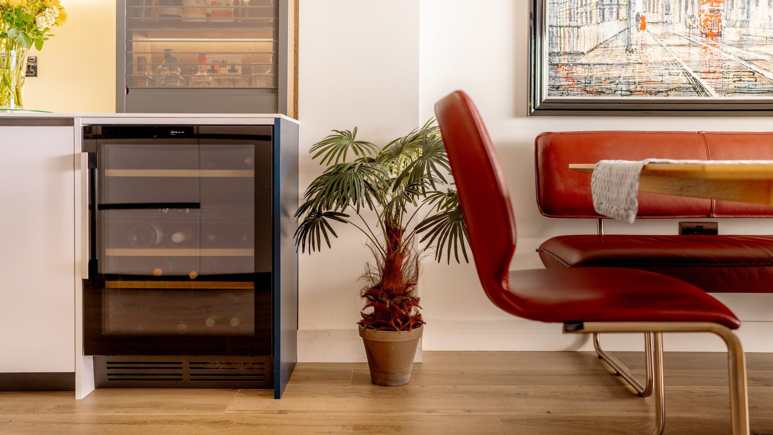 A modern dining area features a red leather chair and bench seat with a wooden dining table. A potted palm sits on the floor next to a small wine fridge integrated into a white cabinet. A framed cityscape painting hangs on the wall above the bench.