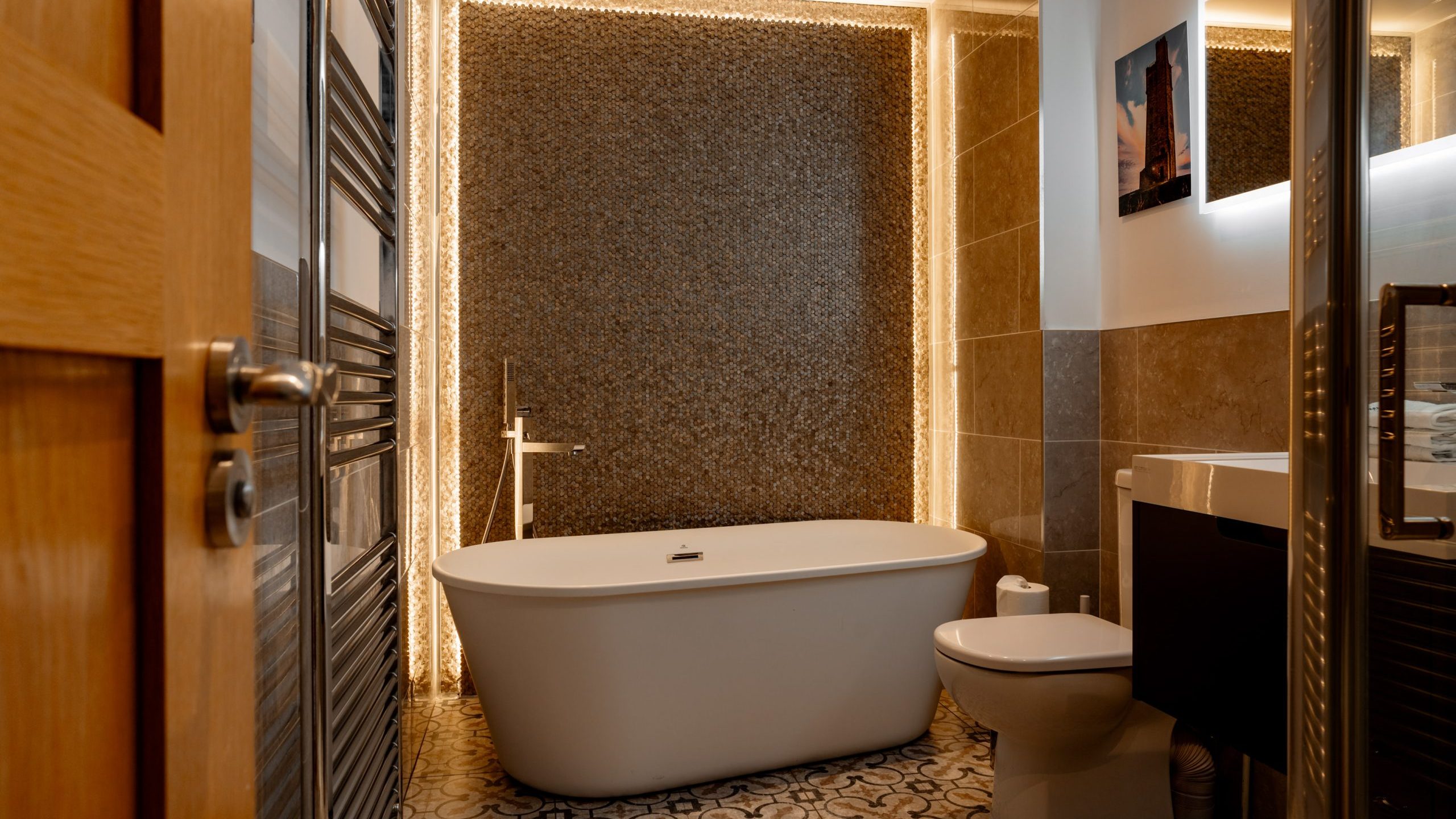 A modern bathroom featuring a freestanding white bathtub against a textured pebble wall with soft backlighting, a toilet, a towel rail, and patterned floor tiles. A black vanity unit with a sink and a mirror is visible alongside a partially open wooden door.