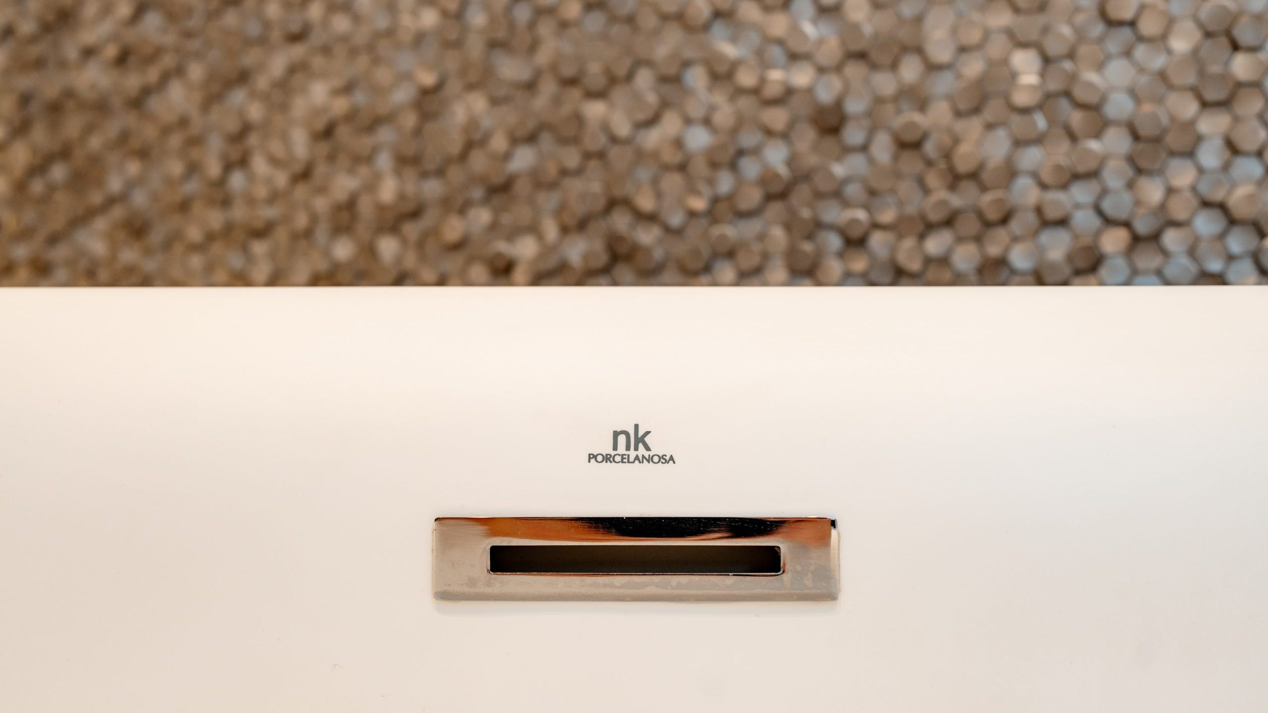 A close-up of a Porcelanosa nk product, featuring a sleek, minimalist design with a small slot, presumably for airflow or drainage. The background consists of a pebble-like texture in shades of gray.