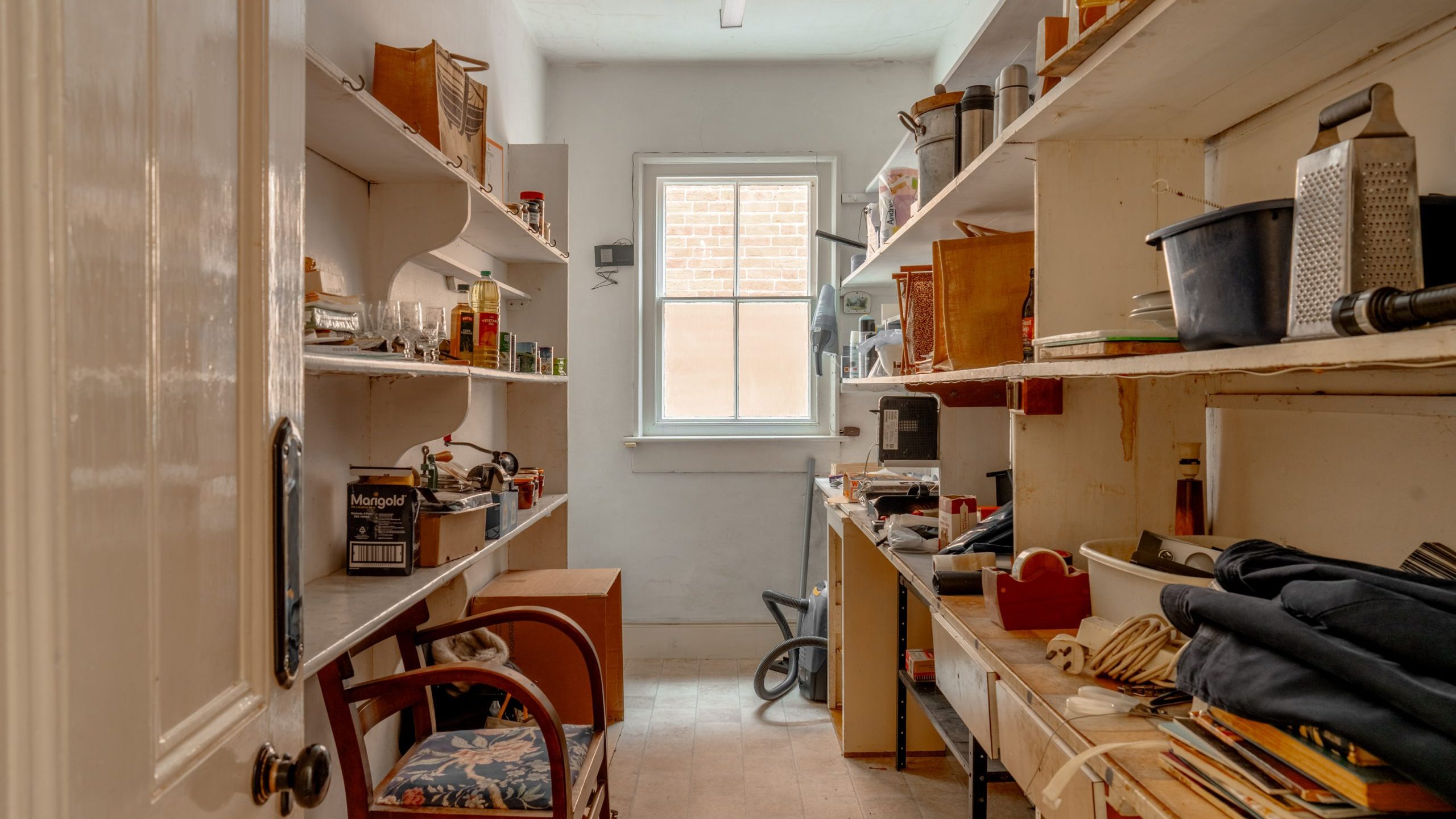 A cluttered, narrow room with shelves on both sides filled with various tools, jars, and miscellaneous items. A window at the far end brings in natural light. The floor is covered with boxes and objects, and a chair is positioned next to a wooden table.