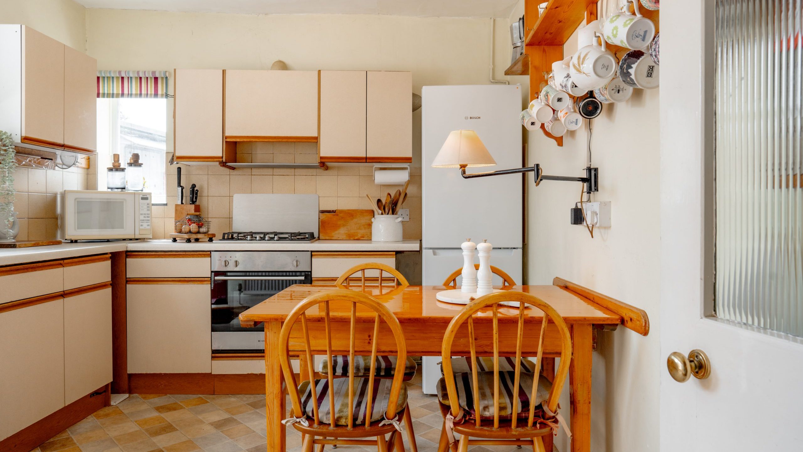 A cozy kitchen with beige cabinets and white appliances, including a fridge and microwave. A small wooden dining table with two chairs sits in the center. A wall-mounted lamp and a shelf holding various mugs are on the right. Natural light streams in from a window.