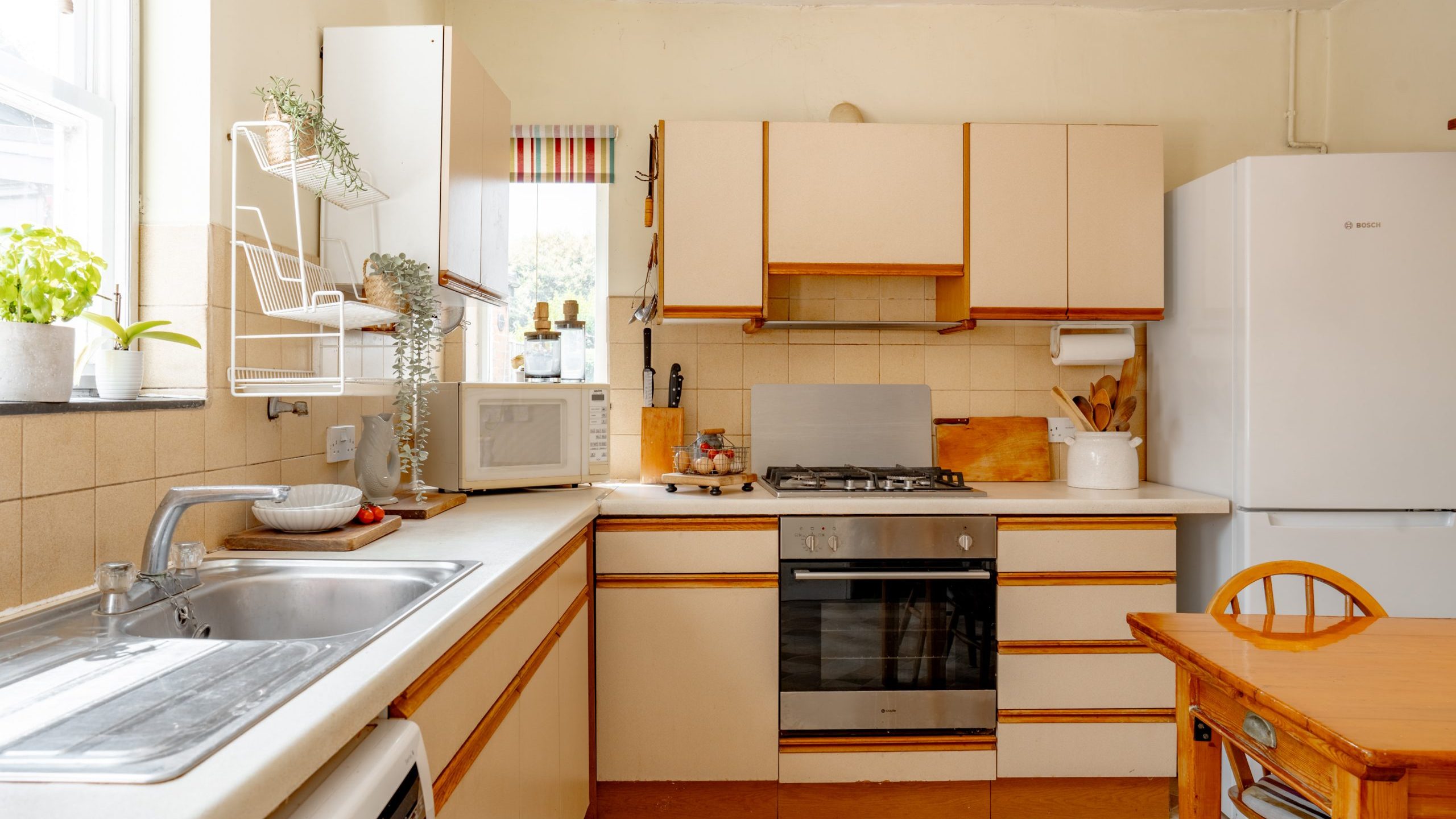 A cozy kitchen with beige cabinetry, a gas stove, an oven, a white refrigerator, and a wooden dining table with chairs. There is a microwave and various kitchen utensils on the countertop, plants on the windowsill, and a double sink under a window.