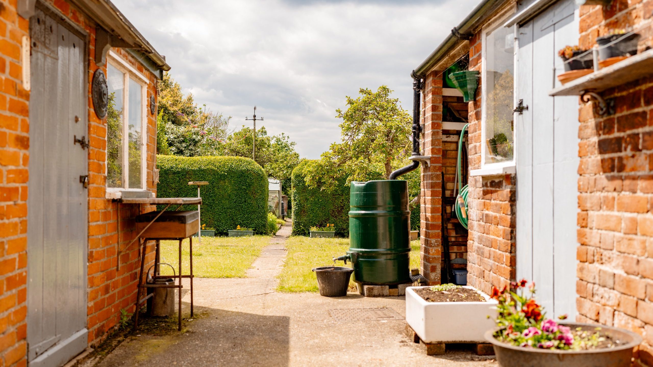A bright outdoor scene featuring a narrow passage between two red brick buildings with gray doors. A green water barrel and potted plants can be seen. The passage leads to a lush garden with a neatly trimmed hedge and trees under a partly cloudy sky.