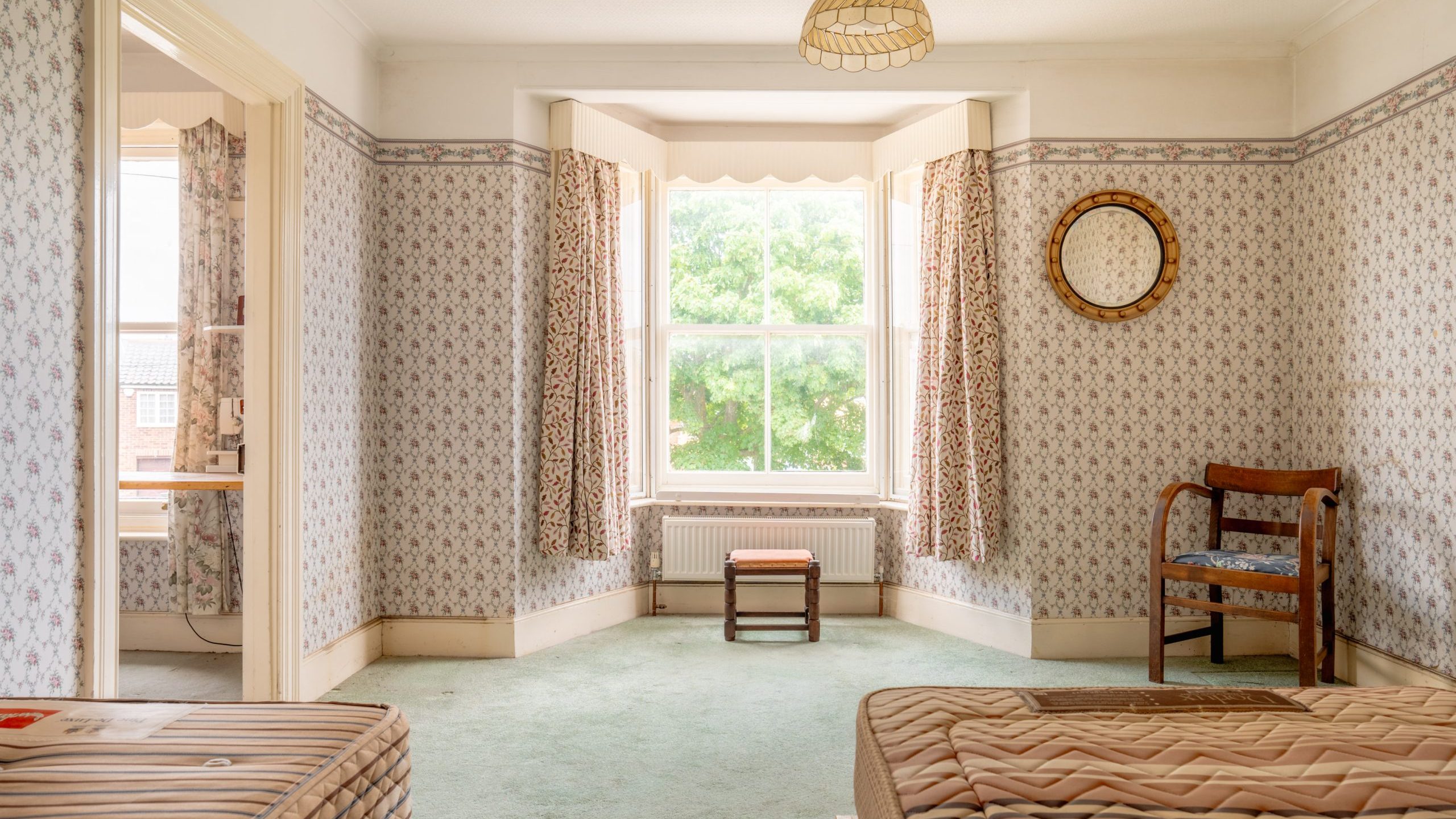 A vintage bedroom with floral-patterned wallpaper, green carpet, and a large bay window with floral curtains. The room features a wooden chair, a small stool, a round wall mirror, and two single beds with mattresses. Natural light fills the space through the window.
