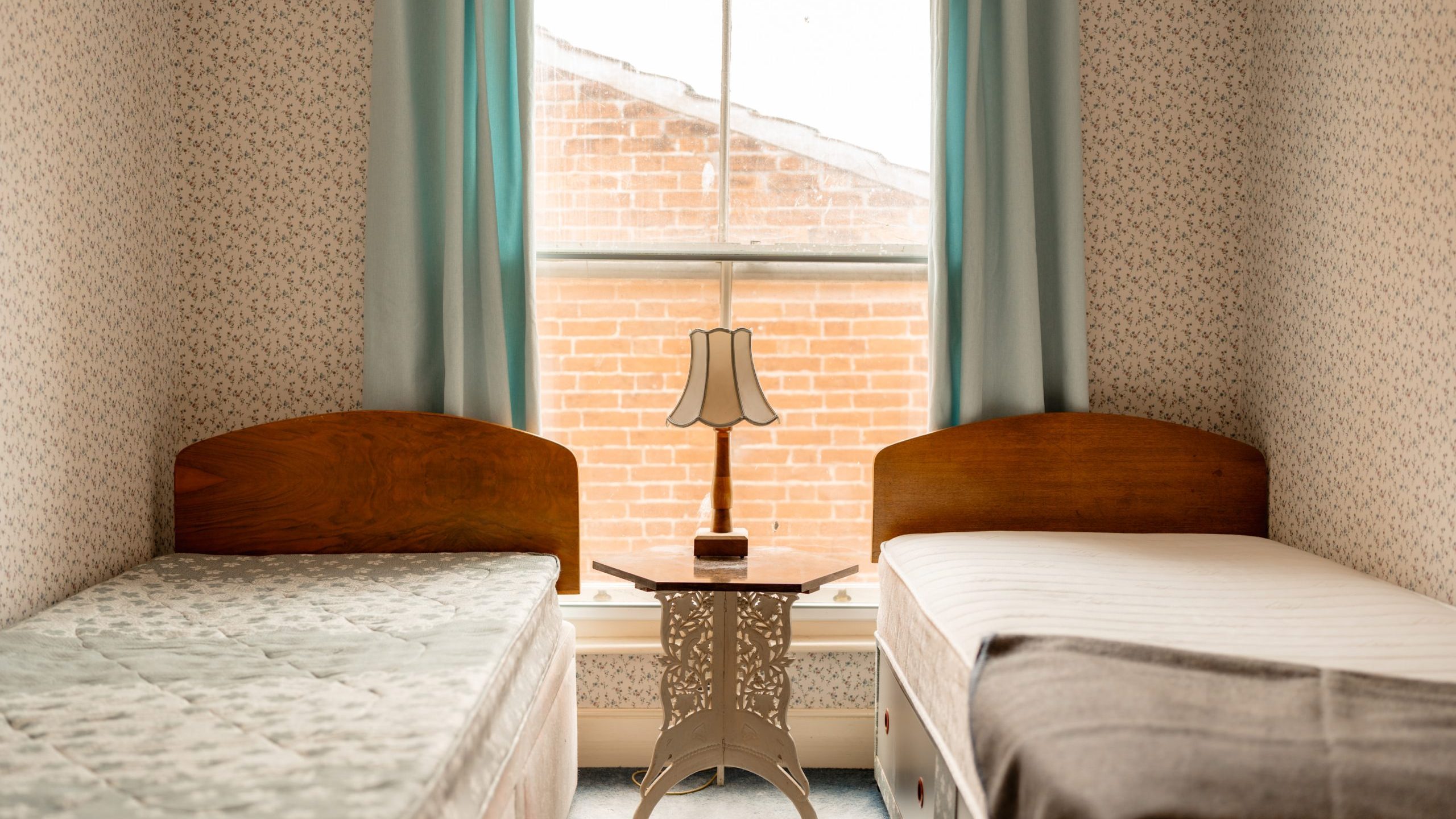 A small bedroom with two single beds placed on either side of a wooden nightstand. The beds have different colored mattresses, one green and one white, with light bedding. A window with light blue curtains and a central table lamp overlooks a brick wall outside.