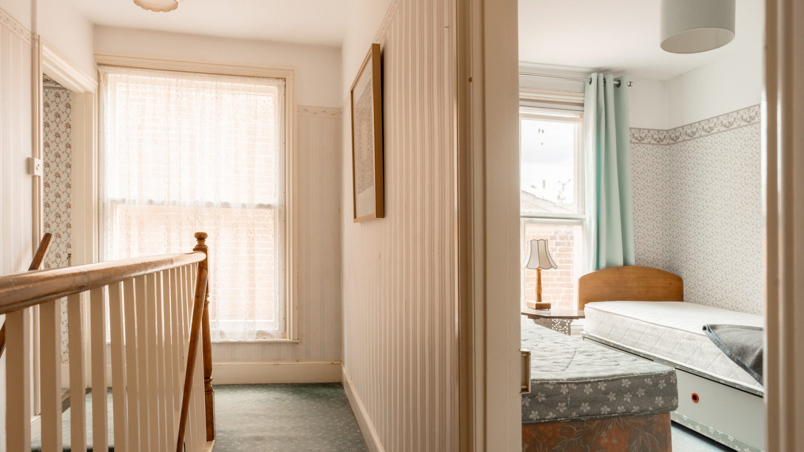 A narrow, sunlit corridor with wooden railings and stairs on the left leads to a bedroom on the right. The bedroom has two beds, a window with sheer curtains, and white walls with a floral border. The space is minimalistic and bright.