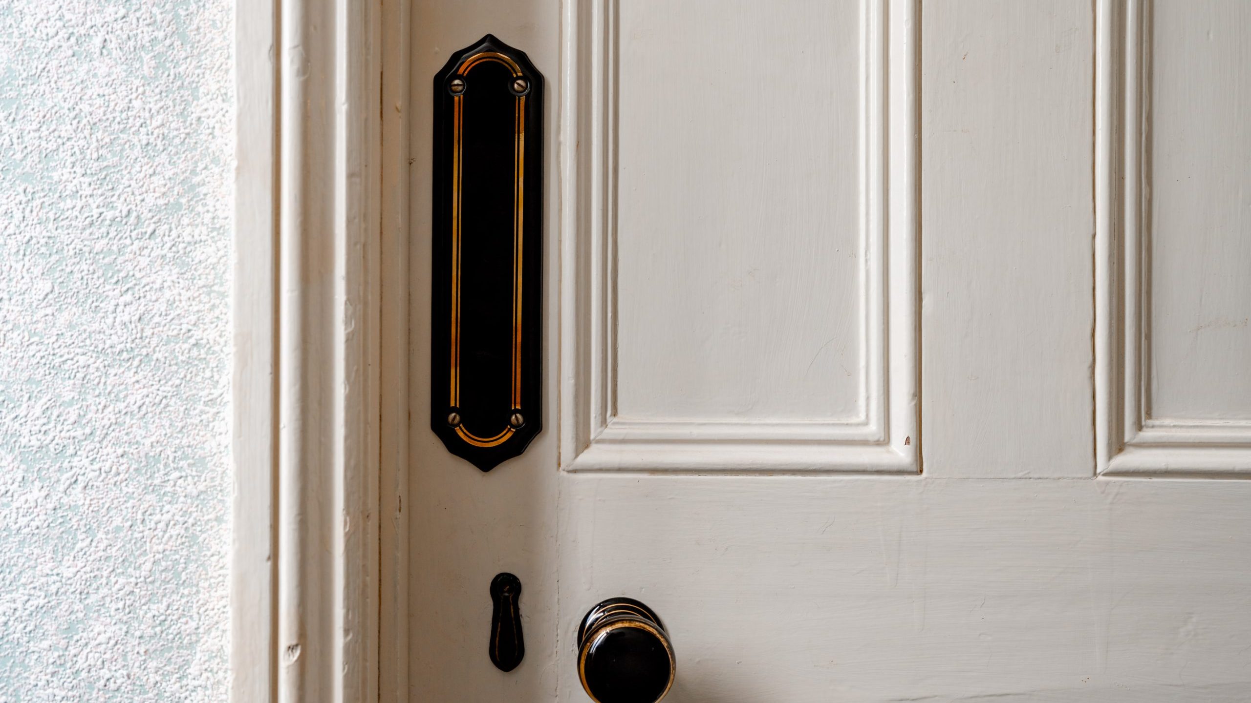 A white wooden door with rectangular paneling features a dark brass door knob and an old-fashioned, black door escutcheon with decorative gold trim. The door is partially ajar, with textured light blue wallpaper visible in the background.