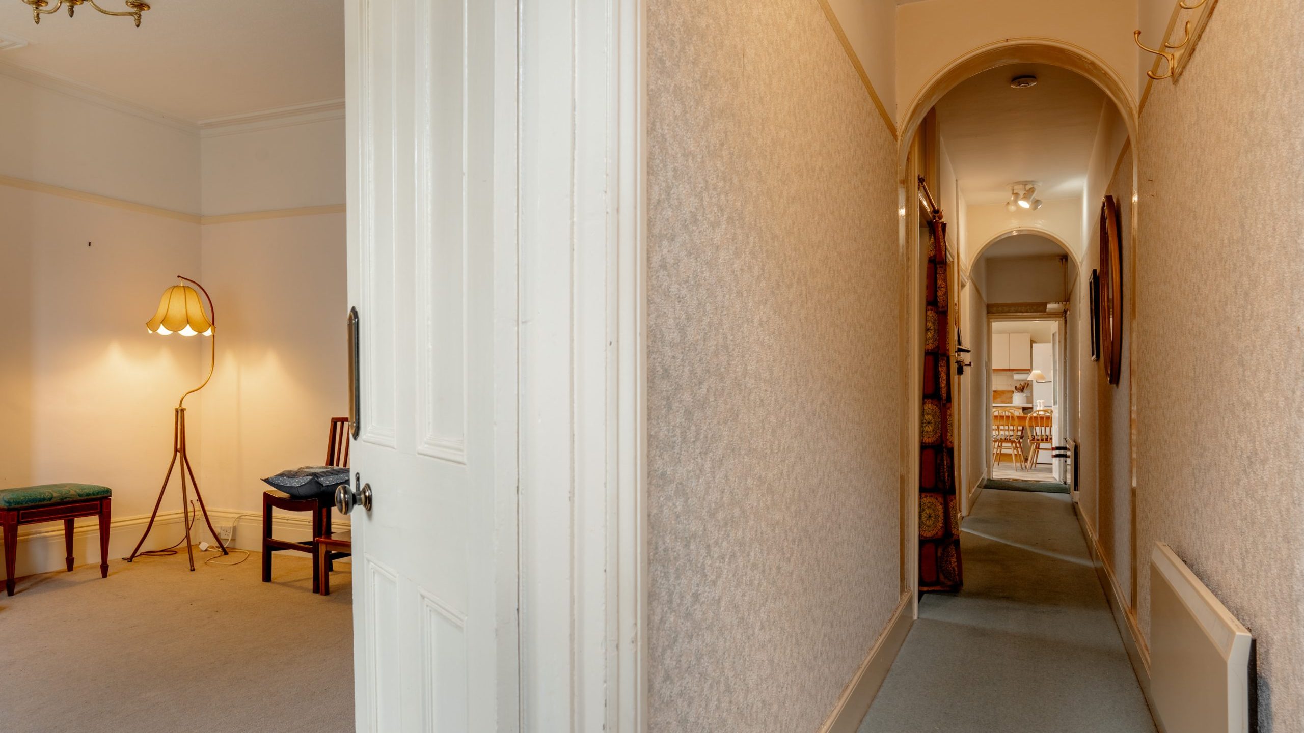 A view of the interior of a house showing a hallway and a living room. The living room has a floor lamp and a wooden chair. The hallway, with an archway, leads to another room with a visible dining table and chairs at the far end. The walls are adorned with wallpaper.
