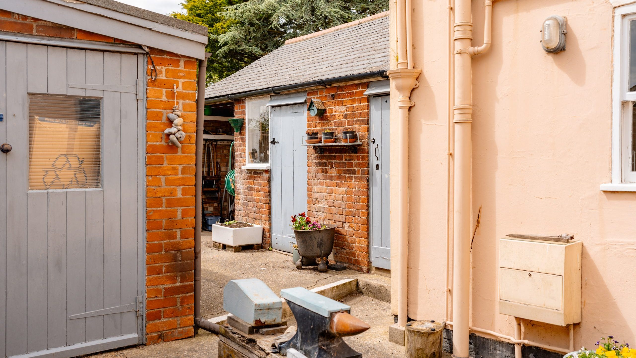 A narrow, outdoor space between pink and brick buildings with a laundry cabinet and pipe on the right. An anvil is visible on the ground. A grey shed with a window stands on the left, and another brick building with two doors and small flower pots extends from the back.