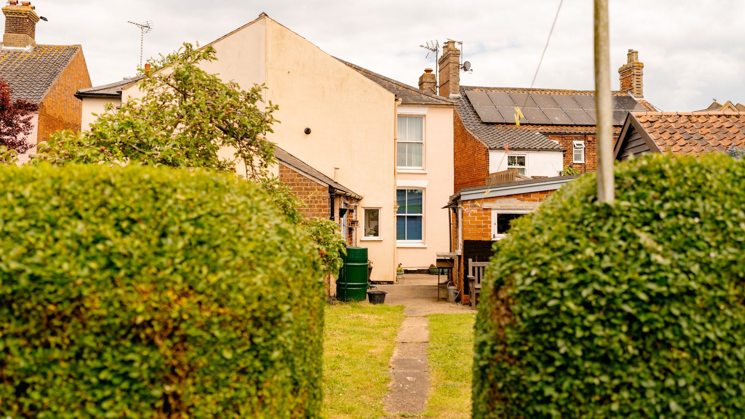 A narrow paved path leads through a well-maintained lawn bordered by two large, neatly trimmed hedges. In the background, there are several connected houses with brick walls and solar panels on the roof. The overcast sky hints at a cloudy day.
