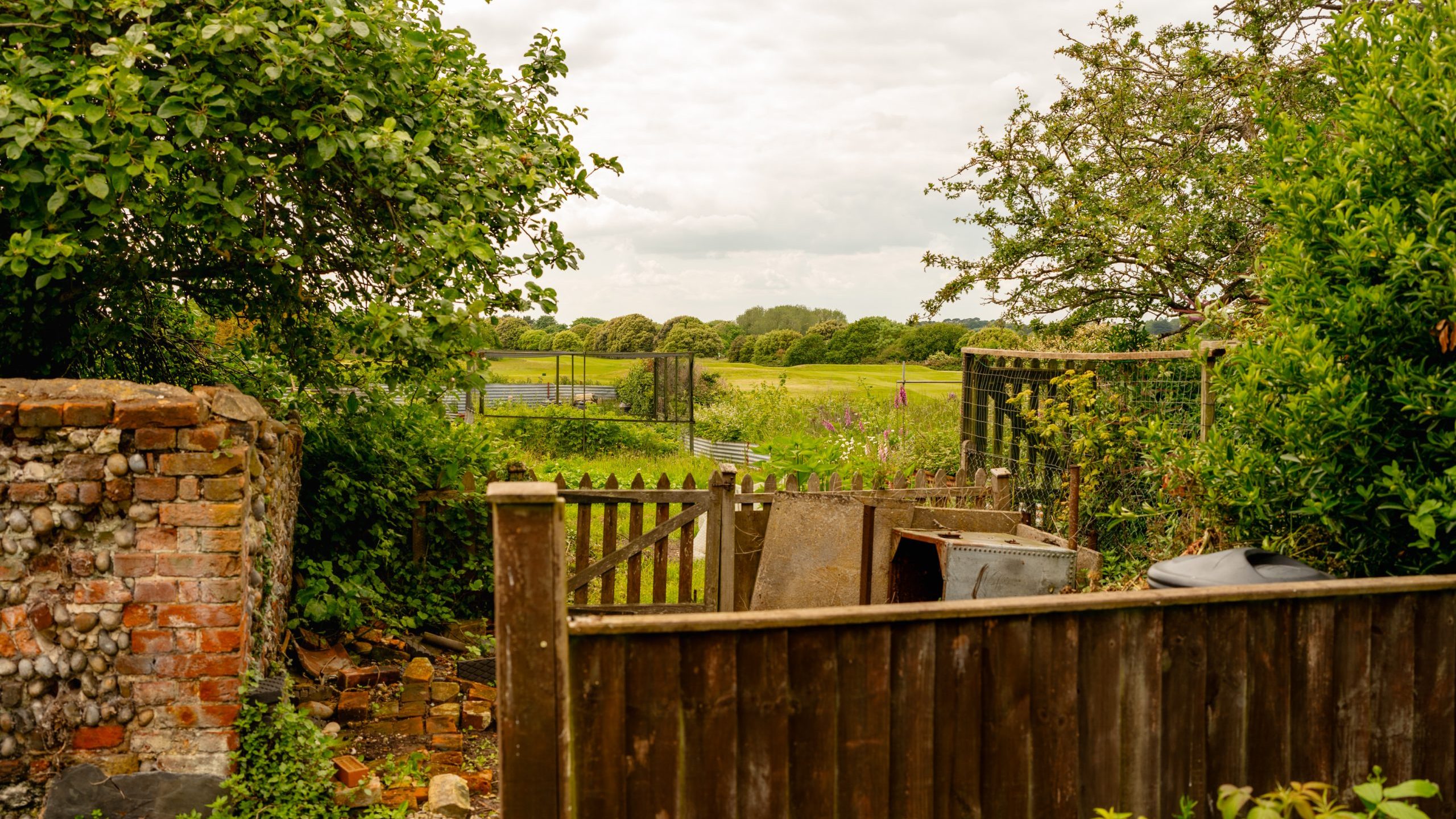 A rustic wooden fence and gate lead to an overgrown garden with various plants and greenery. In the background, a field stretches out under a partly cloudy sky, bordered by trees. A weathered brick wall and wooden structures are also visible.