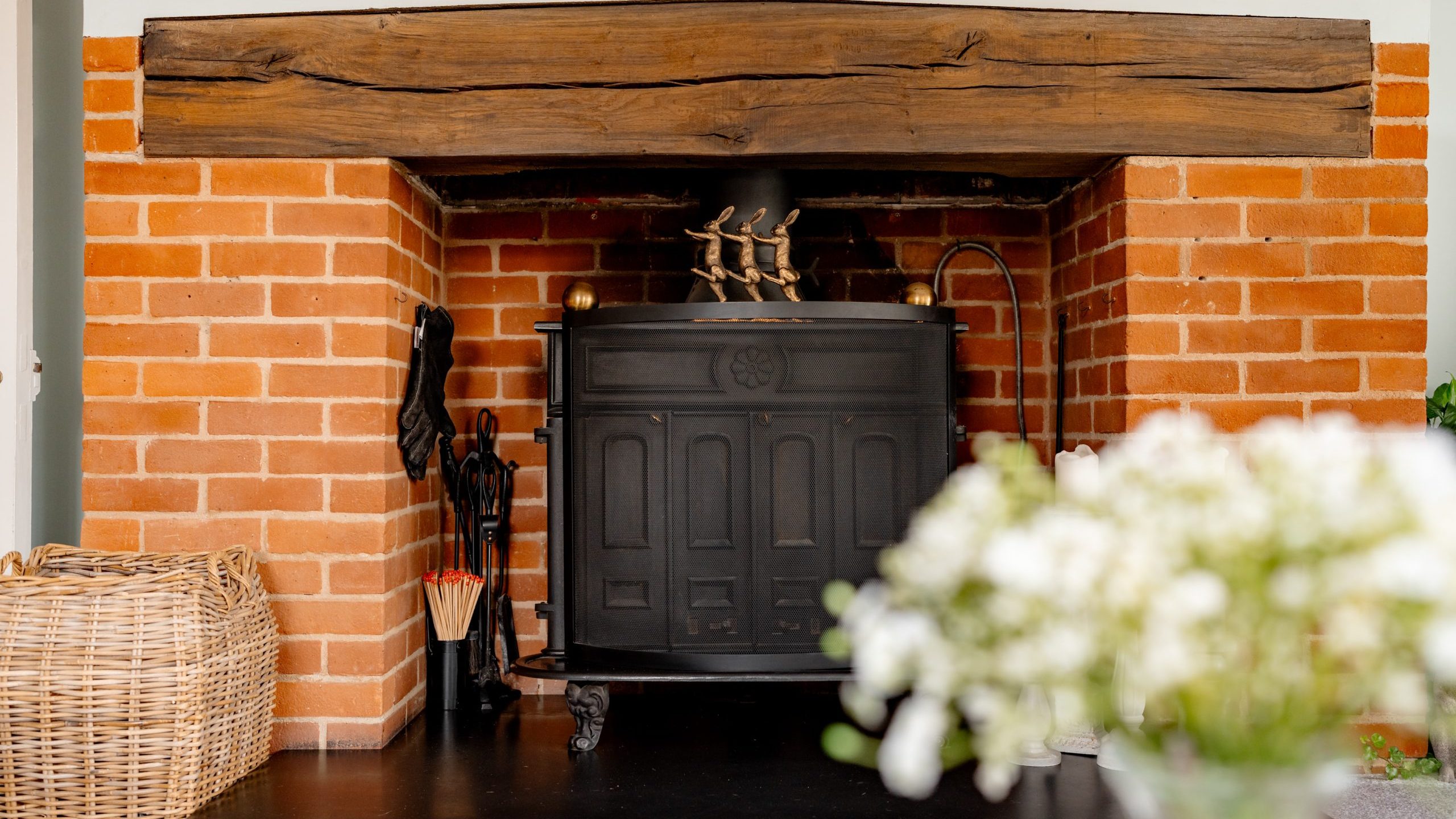 A cozy room featuring a rustic brick fireplace with a dark cast iron stove insert and a wooden mantelpiece. A wicker basket filled with kindling sits to the left, while a bouquet of white flowers is in soft focus in the foreground.