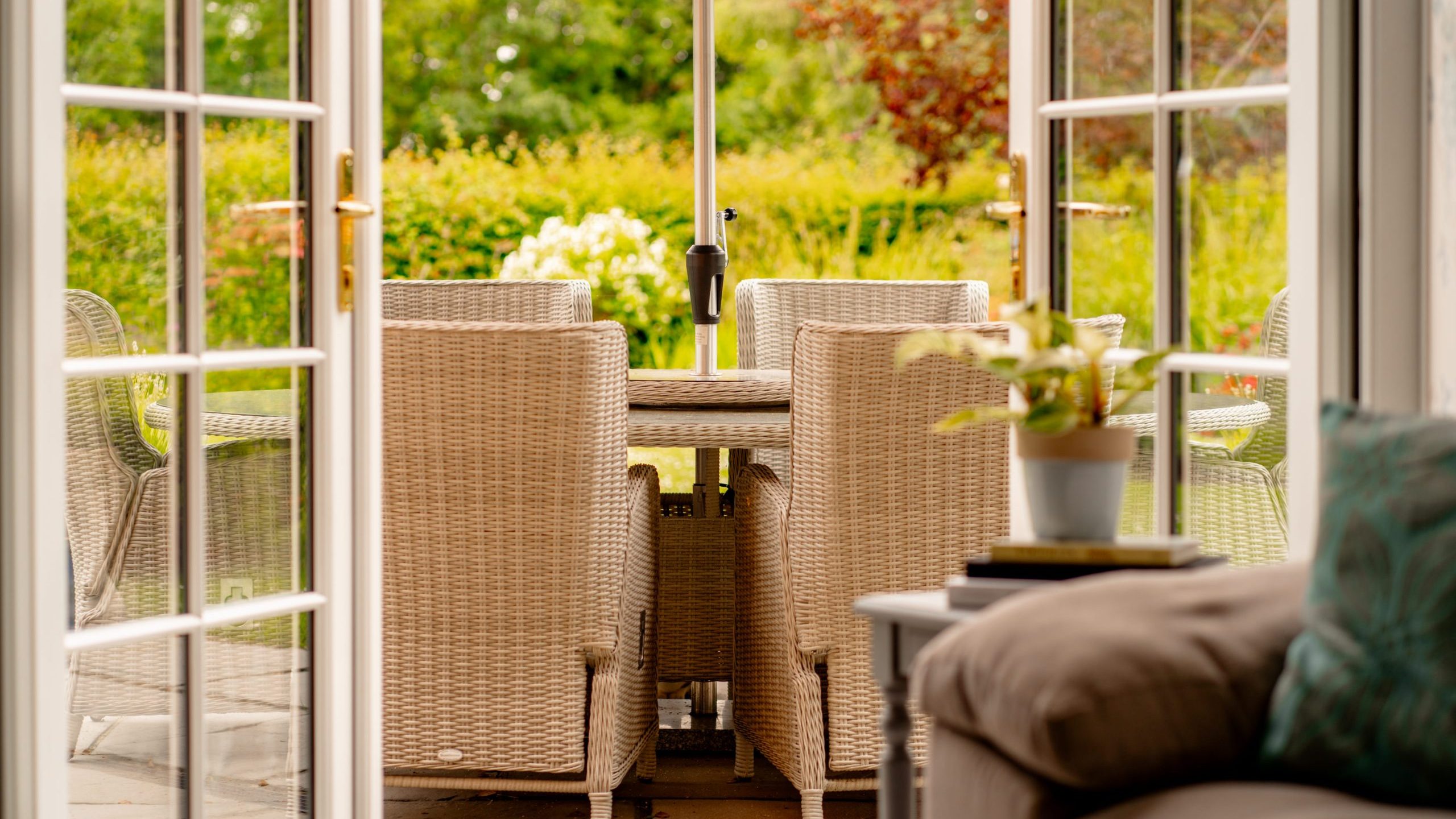 The image shows an inviting patio with wicker chairs and a table set for outdoor dining. The patio doors are open, leading to a lush garden with green foliage and a hedge in the background. A cozy chair with cushions is in the foreground, suggesting a comfortable indoor space.