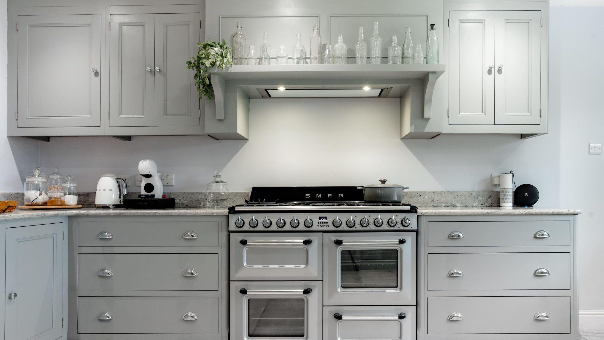 A modern kitchen featuring light gray cabinets and drawers, a countertop with various kitchen appliances, and a stainless steel SMEG oven with multiple compartments. Decorative glass bottles and greenery are on the shelf above the oven.