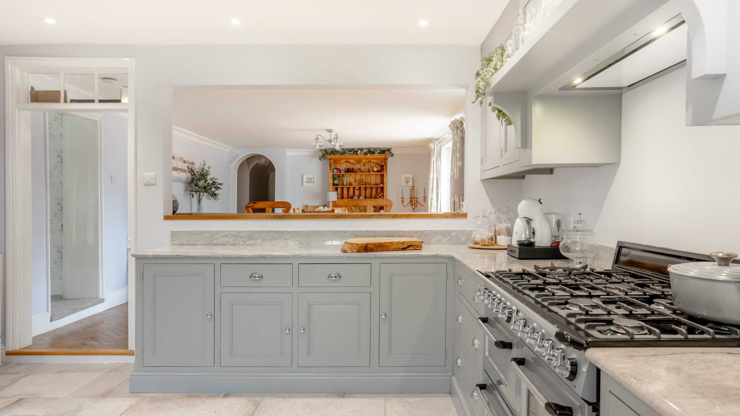 A modern, bright kitchen with light grey cabinets and a granite countertop. A gas stove and oven are on the right side, with cooking utensils and a pot. In the background, a wooden dining table and chairs are visible through an open doorway.