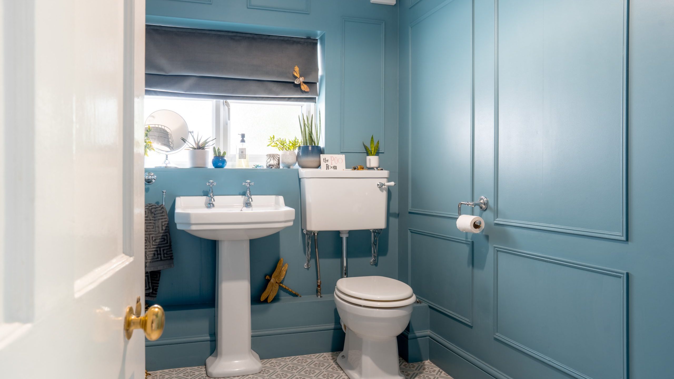 A small, modern bathroom with blue walls and white fixtures. It features a pedestal sink, a toilet with an overhead cistern, and various potted plants on the windowsill. A gray roller blind covers the window, and patterned gray floor tiles add a stylish touch.