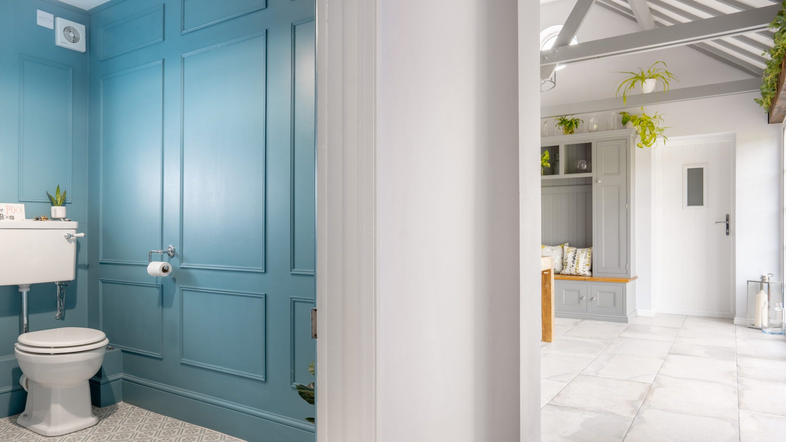 A modern interior features a blue-paneled toilet room on the left, with a white toilet, cistern, and toilet paper holder. The adjacent room to the right has light gray walls, ceramic tiled floor, a bench with cushions, and various potted plants adding greenery.