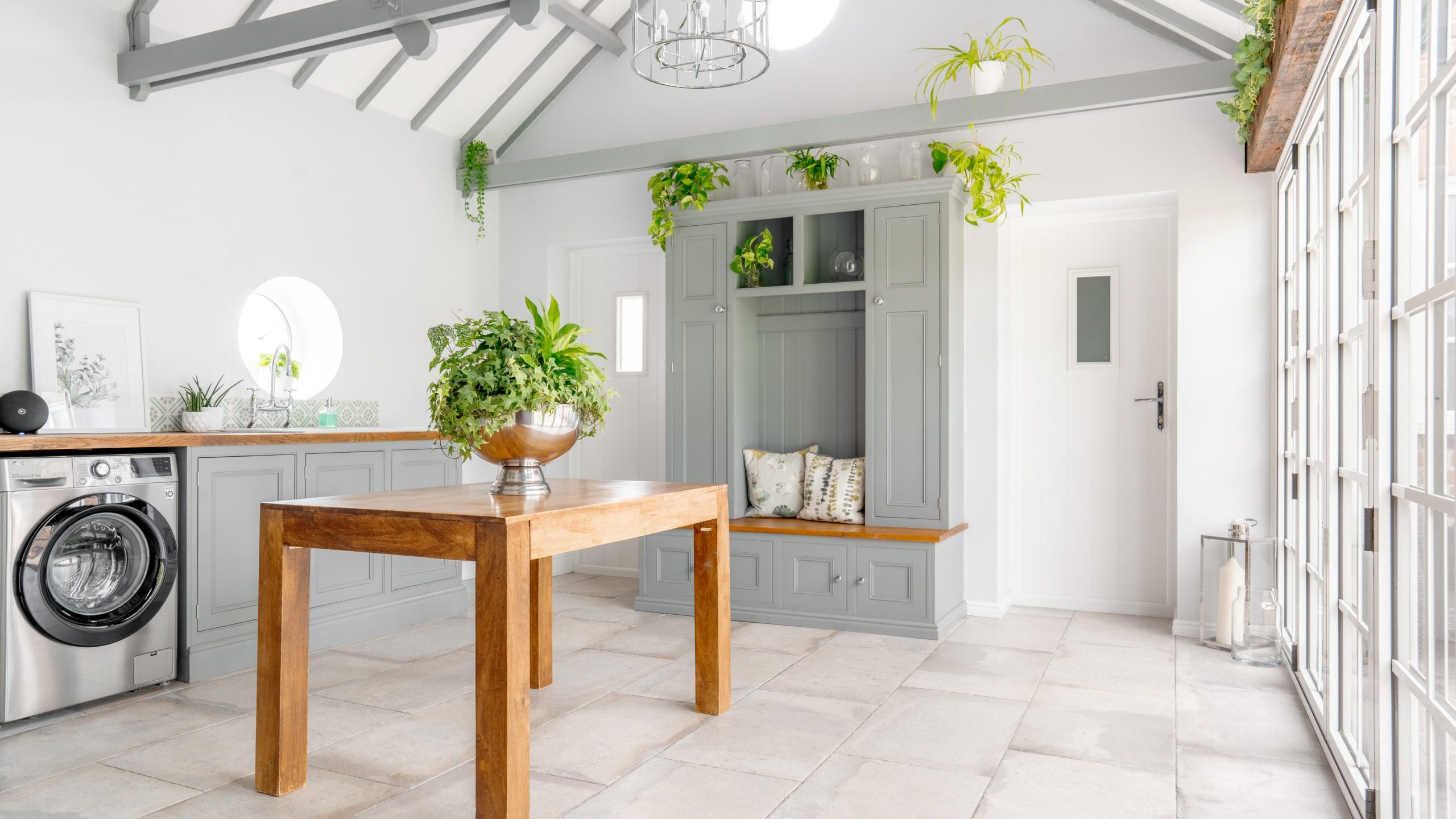 A bright, modern laundry room with a gray, vaulted ceiling, and large windows. A wooden table with a plant centerpiece sits in the middle, a washing machine is on the left. Cabinets, shelves, and greenery decor add charm. A doorway is visible at the back.