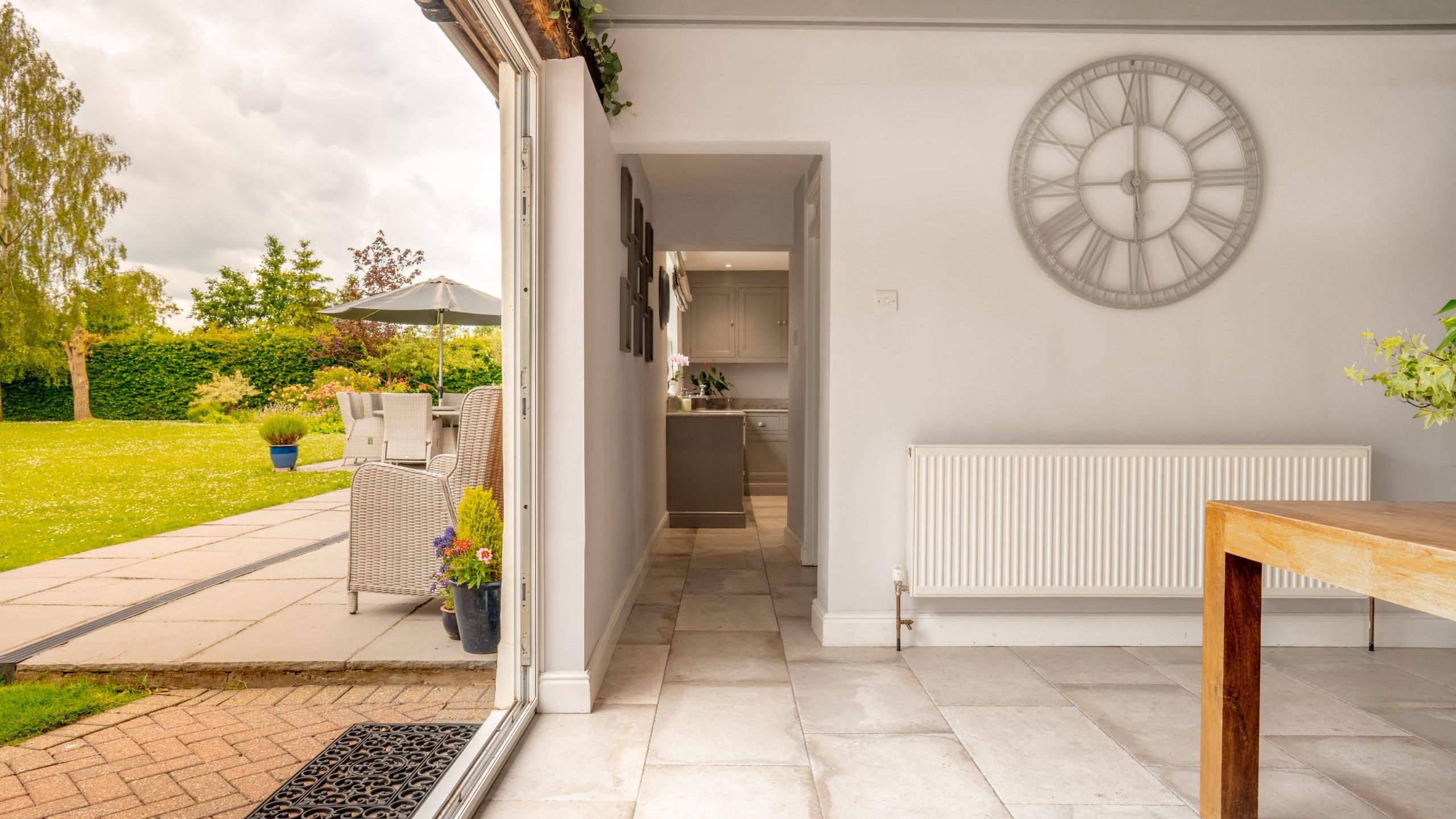 A bright, modern interior space featuring a large wall clock and minimalist furniture. The room opens to a well-manicured garden and patio area with outdoor seating. The scene is filled with natural light from the open sliding doors.
