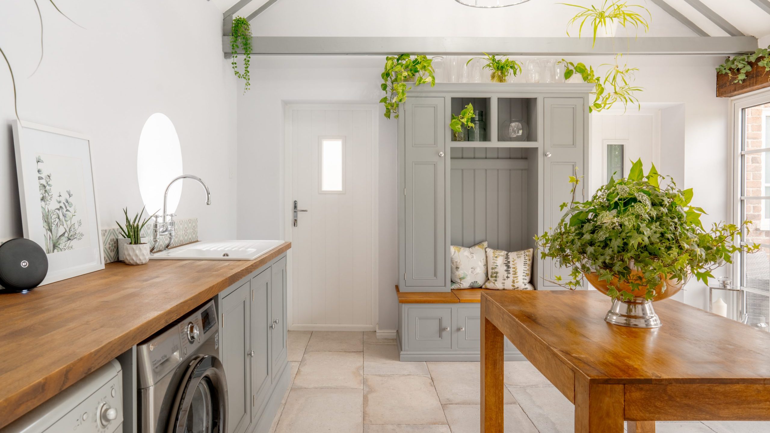 A bright, modern kitchen with white walls and gray cabinetry. There is a wooden countertop with a sink and washing machine. A wooden table with a large plant sits in the center. The room features a high ceiling with exposed beams and greenery accents.