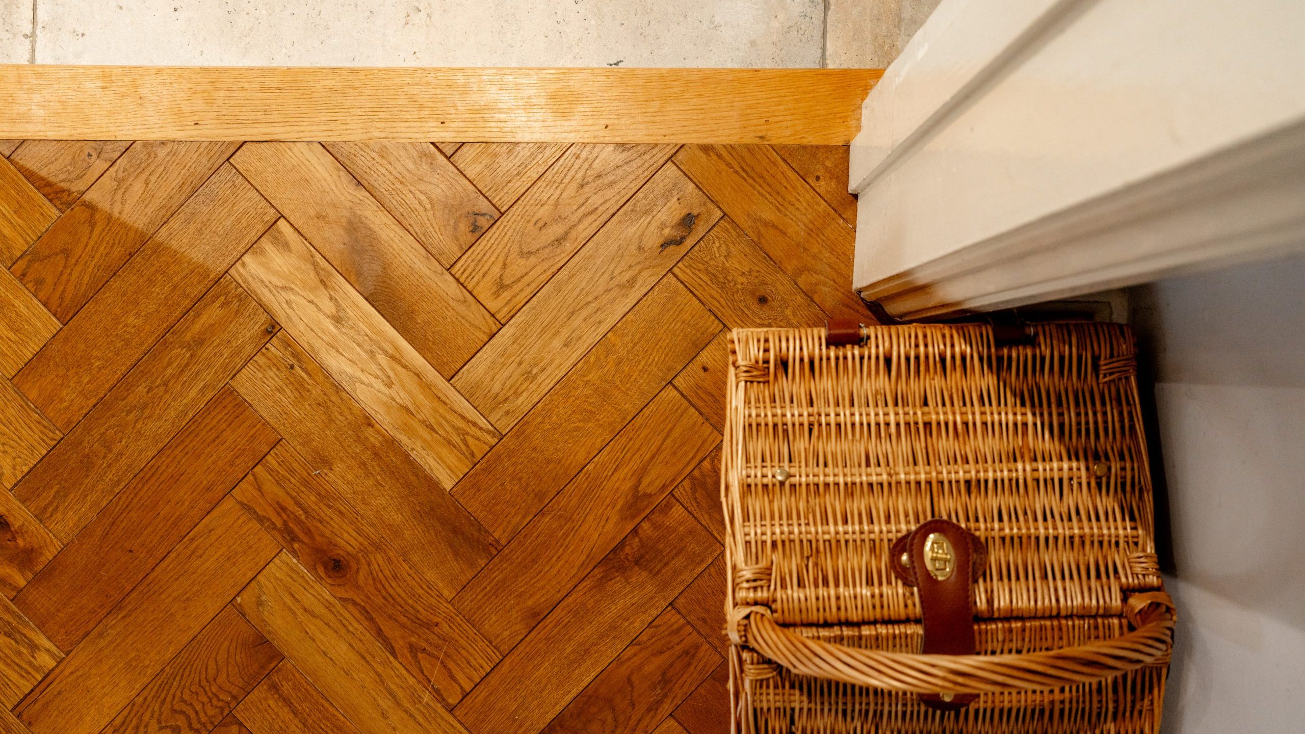 A wicker basket with leather straps sits on a wood floor with a herringbone pattern, adjacent to a light-colored wall and a corner of a white doorframe. The intricate wood design contrasts with the simple basket weave.