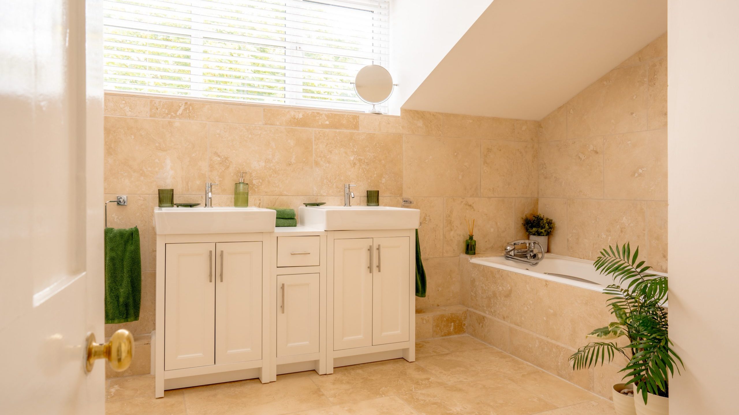 A bathroom with beige tiles, a white double sink vanity with green accessories, a bathtub on the right side, a potted plant beside the bathtub, and a window with blinds above the sinks. There are green towels hanging next to the sinks.