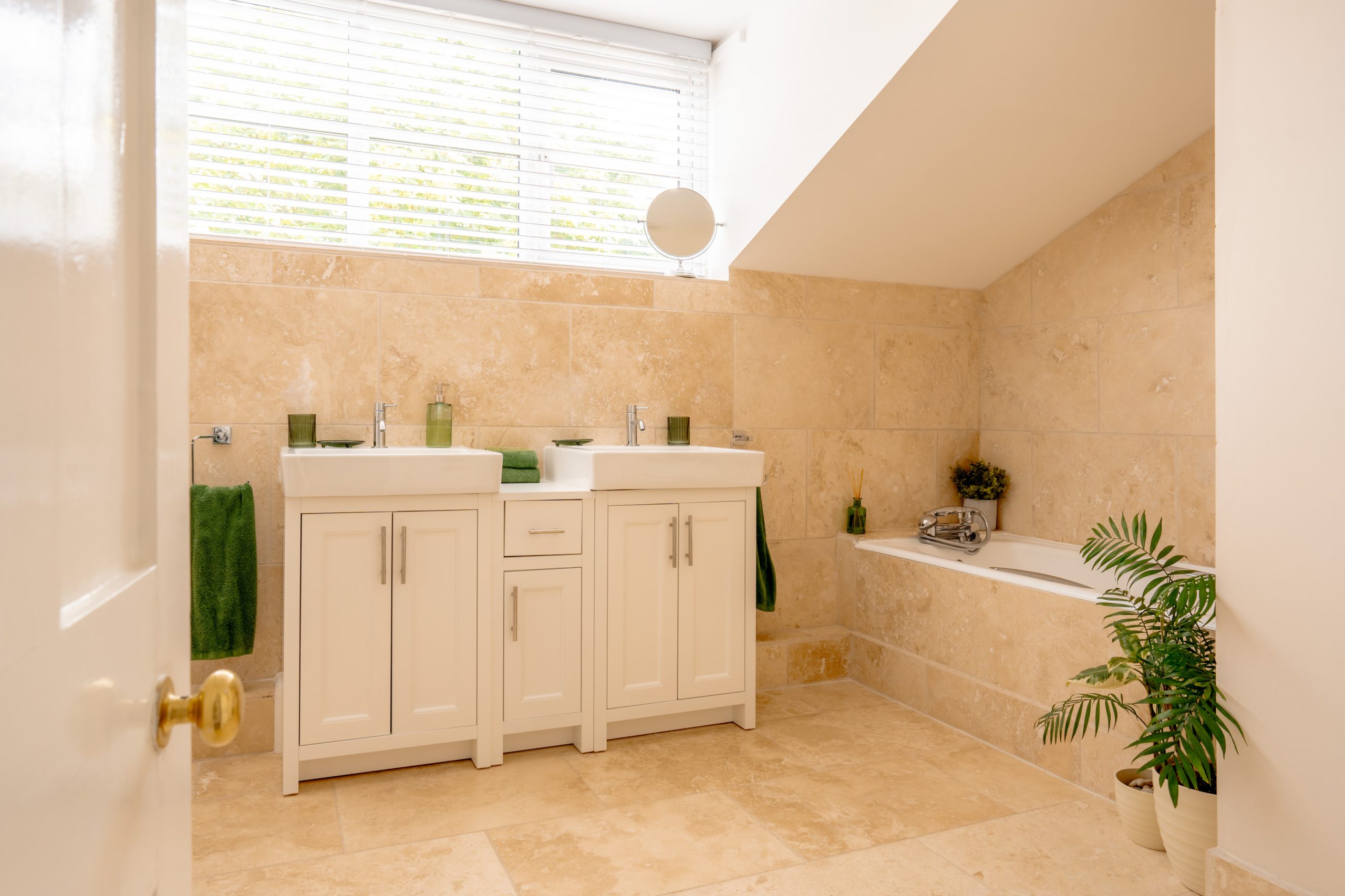 A bathroom with beige tiles, a white double sink vanity with green accessories, a bathtub on the right side, a potted plant beside the bathtub, and a window with blinds above the sinks. There are green towels hanging next to the sinks.
