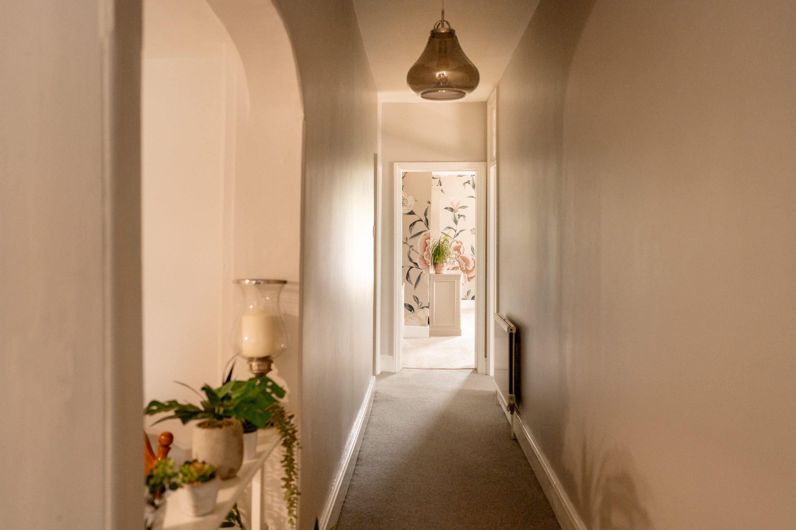 A long, narrow hallway with beige walls and a carpeted floor leads to a well-lit room at the end. A small shelf on the left wall holds plants and a candle. The room at the end has floral wallpaper and more plants, adding a bright and airy feel.