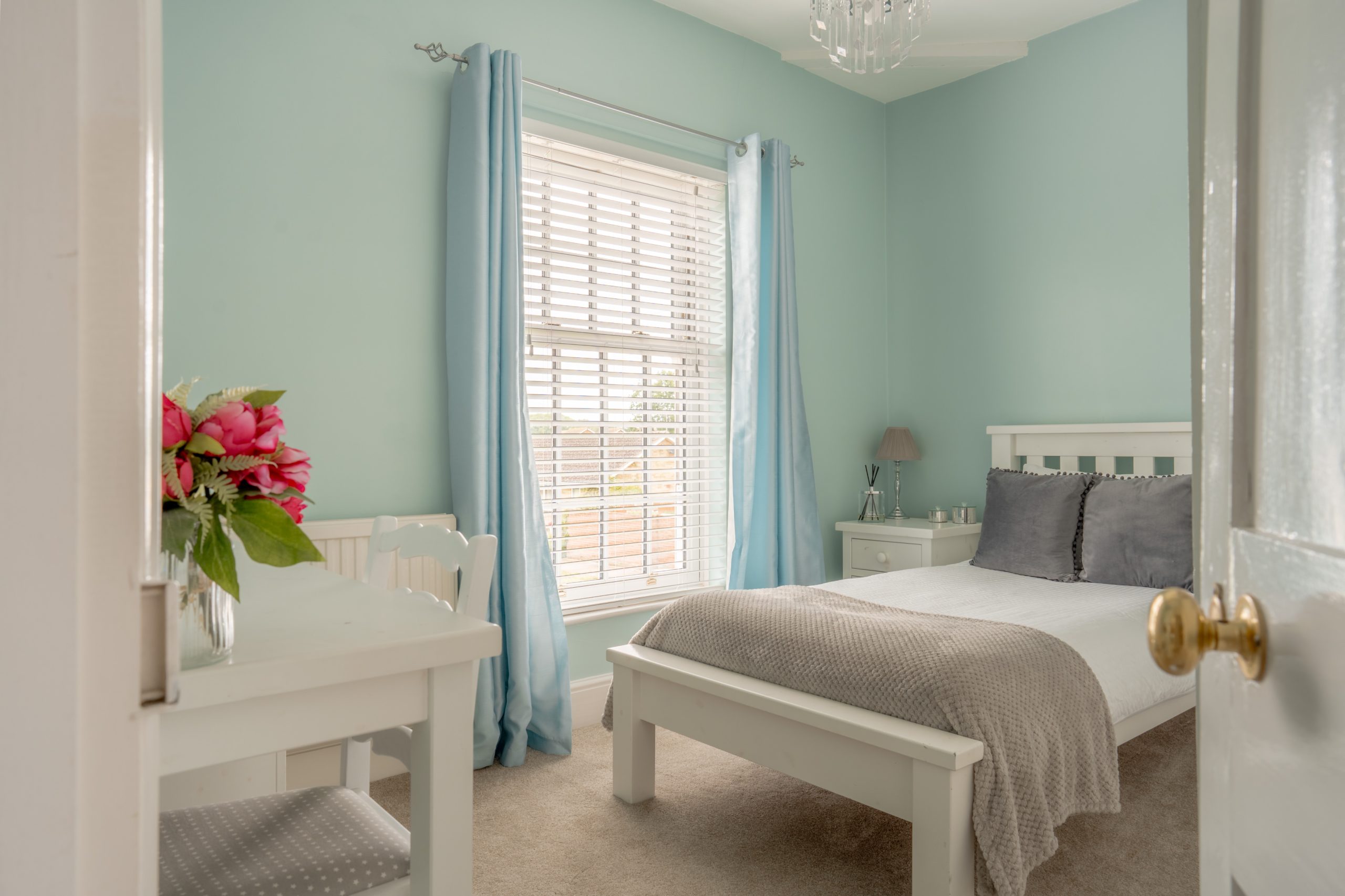 A cozy bedroom with light blue walls and a white bed frame. The bed is made with white and grey bedding. A window with white blinds and blue curtains allows natural light in. A small desk with flowers in a vase is next to the window. A nightstand with a lamp is beside the bed.