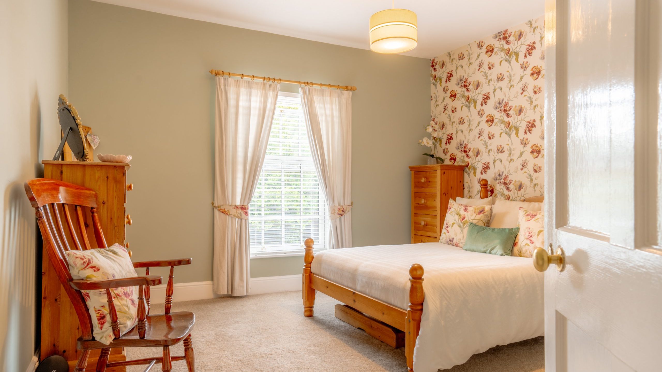 A cozy bedroom with light green walls and a floral accent wall. The room features a wooden bed with white bedding, a rocking chair with floral cushions, and a wooden chest of drawers. Light streams through a window with sheer, white curtains. A ceiling light hangs above.