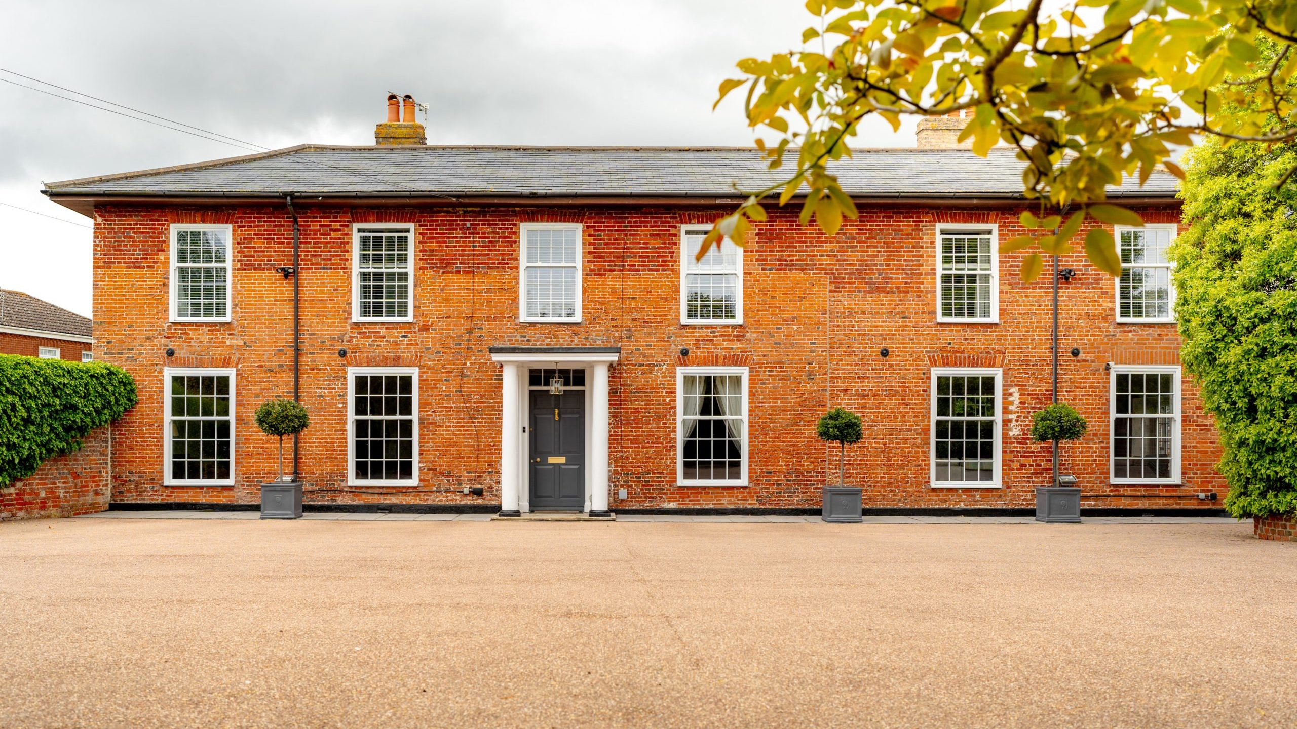 A large, two-story brick building with white-trimmed windows and a central grey door stands under a cloudy sky. Symmetrical potted plants line the front facade, and a manicured tree branch partially frames the right side. A wide, open driveway leads up to the entrance.