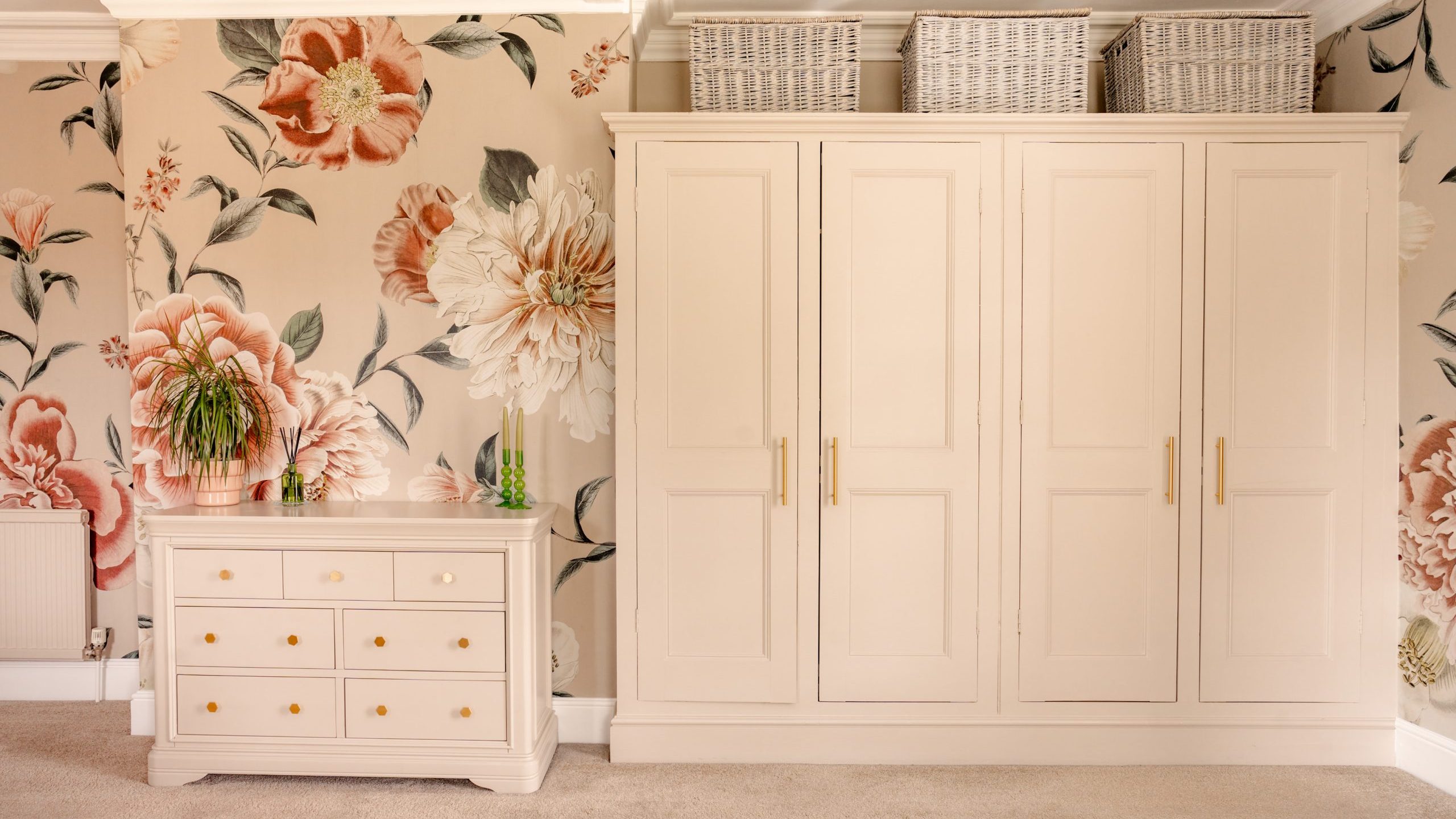 A bedroom corner showcasing a beige dresser with gold drawer knobs, a multi-door wardrobe, and decorative white woven baskets on top. The wall is adorned with a large floral print featuring pink and white flowers. A small green plant is placed on the dresser.