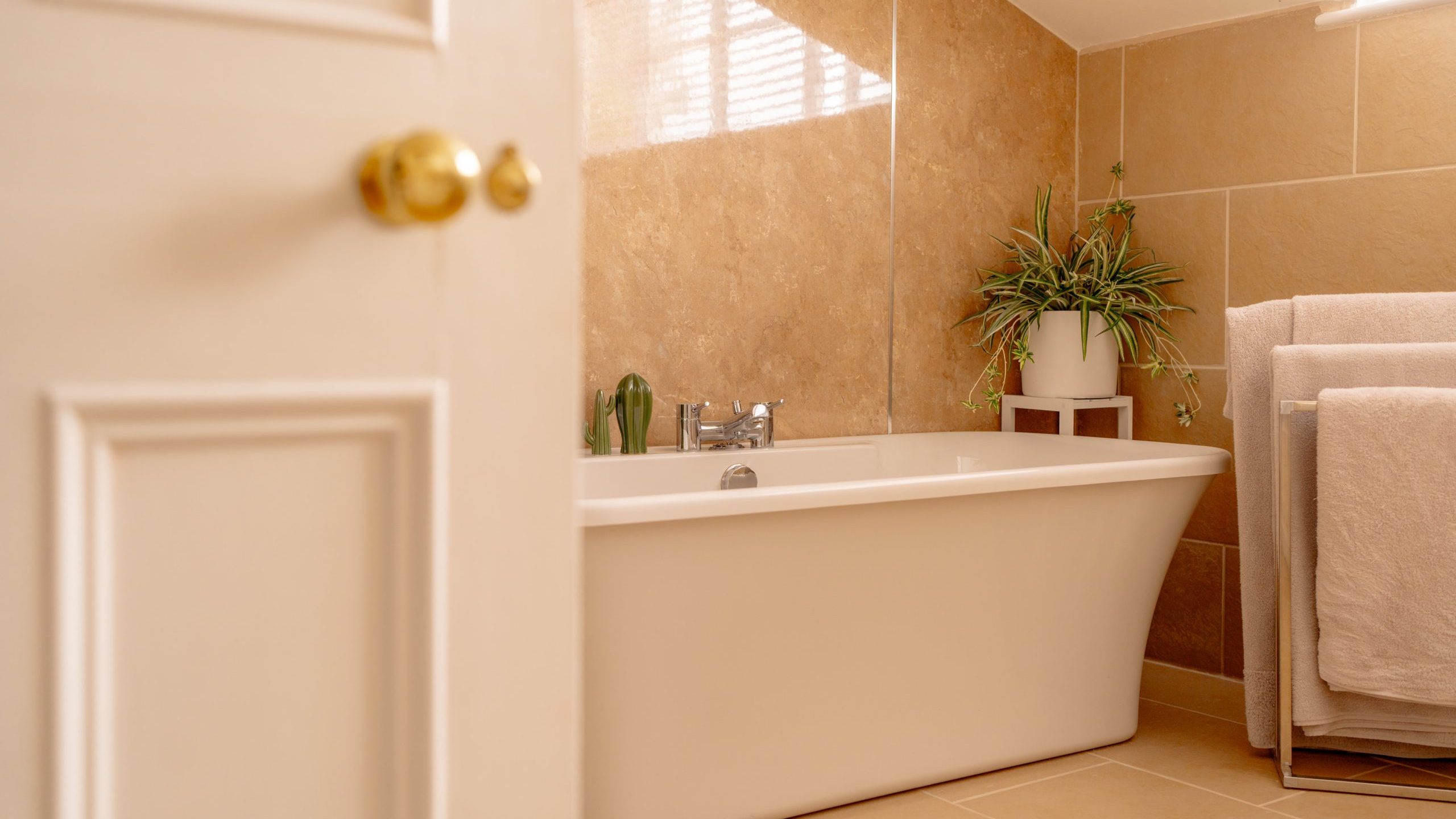 A cozy bathroom with beige tiled walls and floor features a white freestanding bathtub. On the bathtub's edge, a potted green plant adds a touch of nature. Light streams through a window with blinds, illuminating the space. A towel rack with beige towels is nearby.