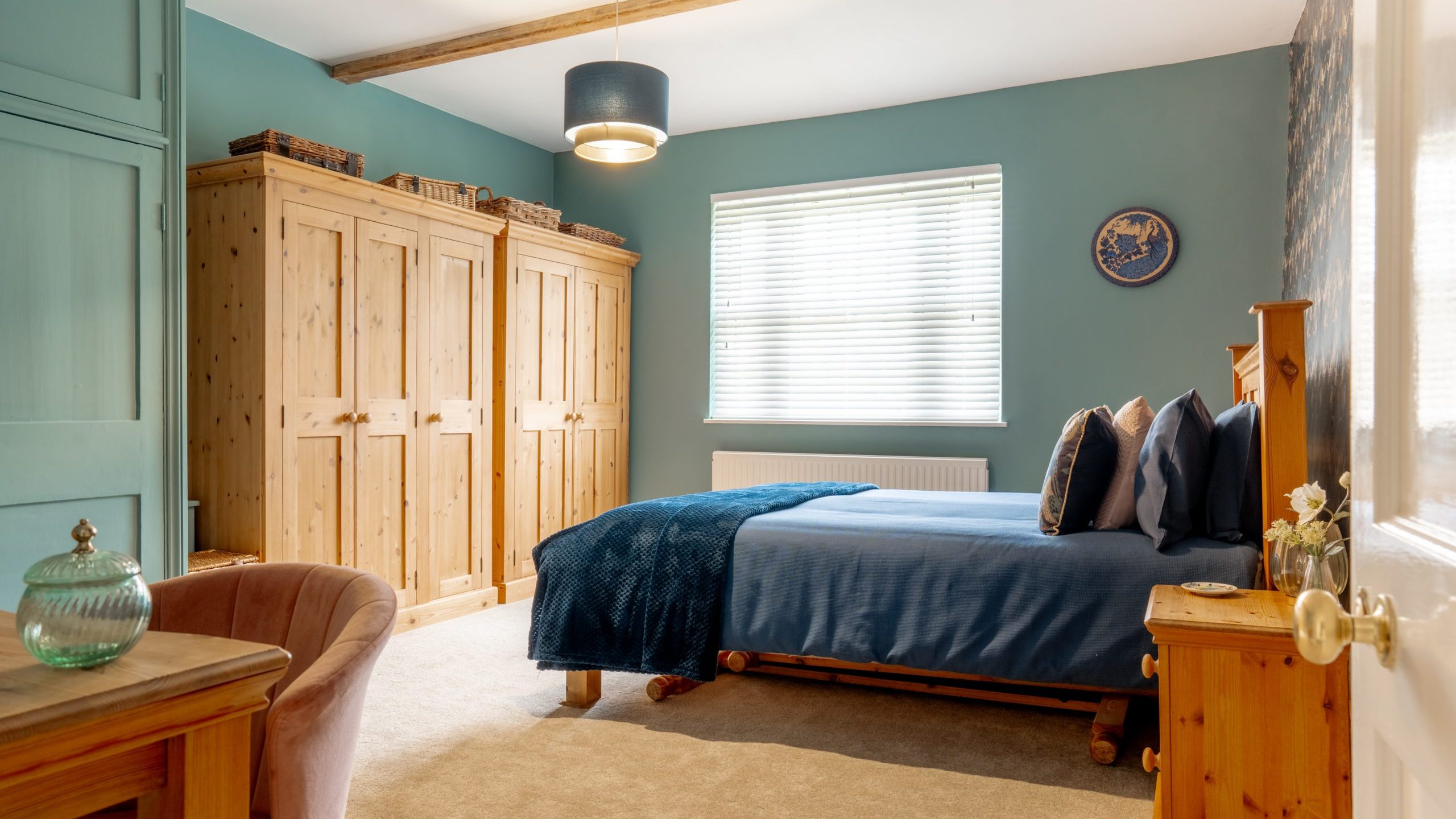 A cozy bedroom with a double bed adorned with blue bedding. Wooden wardrobes line one wall, and a window with blinds allows light to filter in. There's a wooden bedside table with a decorative jar and flowers, and a soft armchair in one corner.