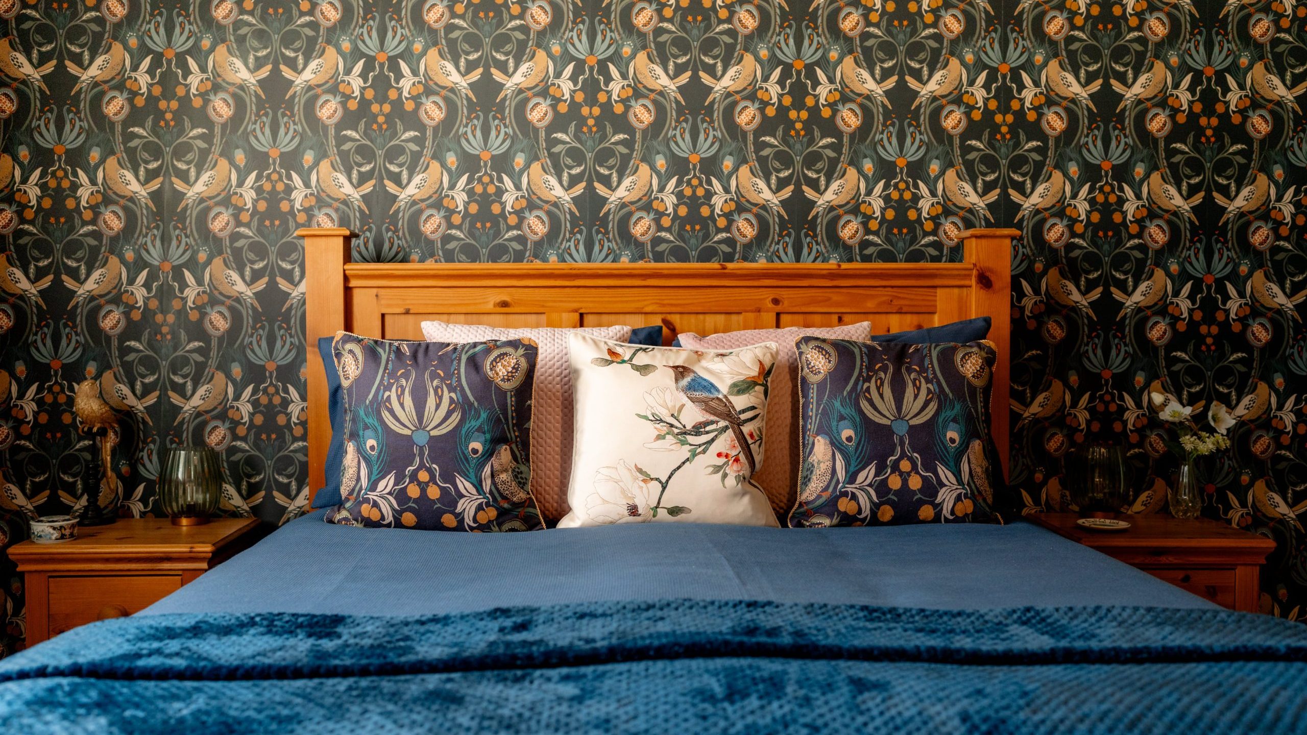 A cozy bedroom featuring a wooden headboard, a blue bedspread with matching blue decorative pillows, and a central pillow with a bird design. The backdrop is an intricate, nature-themed wallpaper with floral and leaf patterns in dark tones.