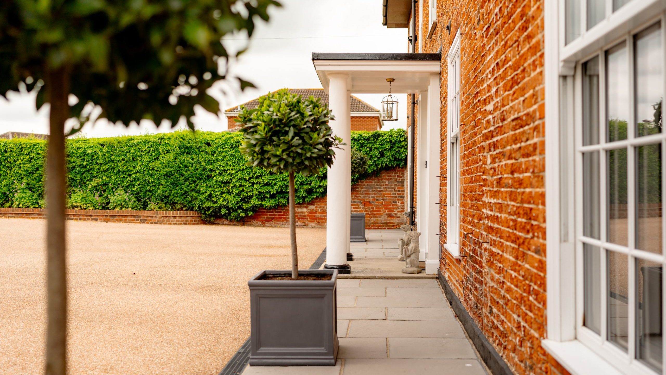 A perspective view of a red brick house with a covered entrance supported by white columns. Topiary trees in square pots line the stone path leading to the entrance. A hedge and brick wall are visible in the background, enclosing a gravel driveway.