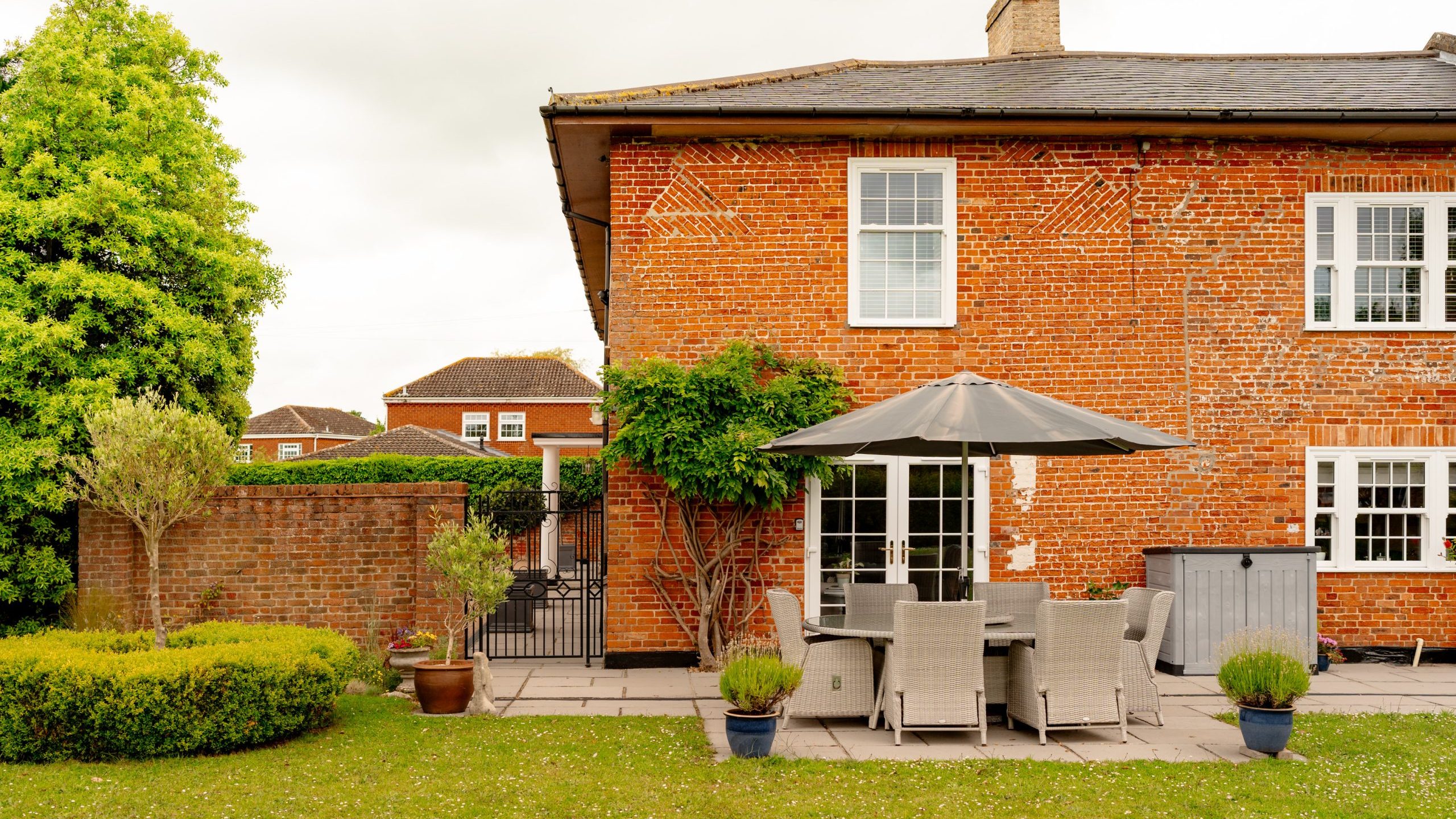 A well-maintained red-brick house with a patio set featuring a table, six chairs, and a large umbrella. The backyard includes green lawn, potted plants, and a trimmed hedge. A tall tree is on the left, and two smaller trees are near the patio. The sky is cloudy.