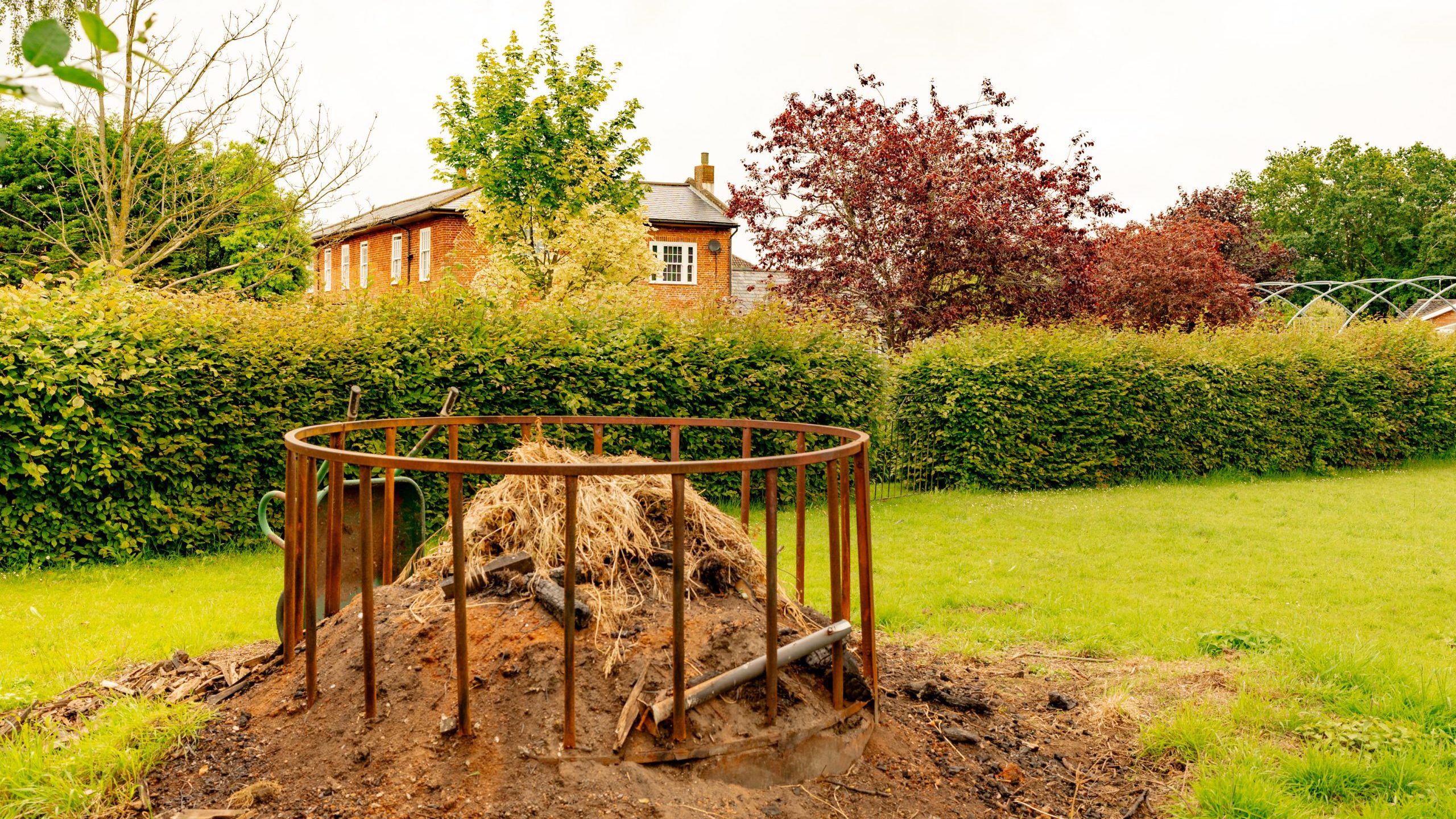 A pile of dry grass and dirt encased in a round metal frame stands in the middle of a grassy area. Behind it is a dense, green hedge, with trees in autumn colors and a brick house partially visible. The sky above is cloudy.