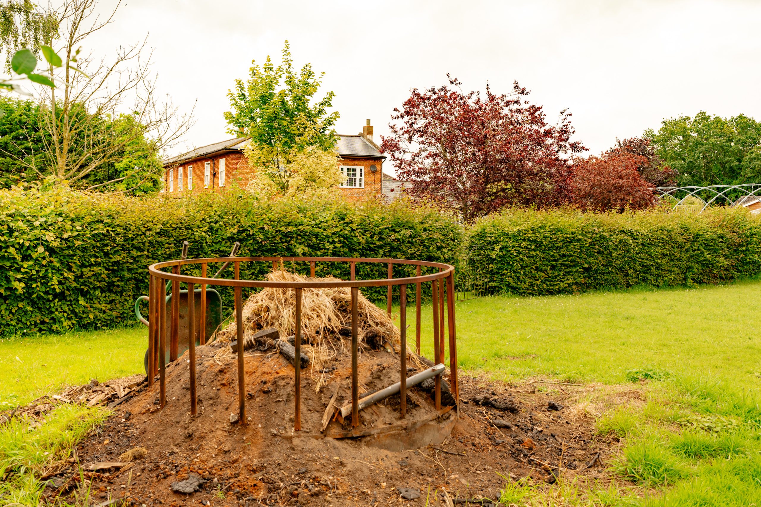 A pile of dry grass and dirt encased in a round metal frame stands in the middle of a grassy area. Behind it is a dense, green hedge, with trees in autumn colors and a brick house partially visible. The sky above is cloudy.