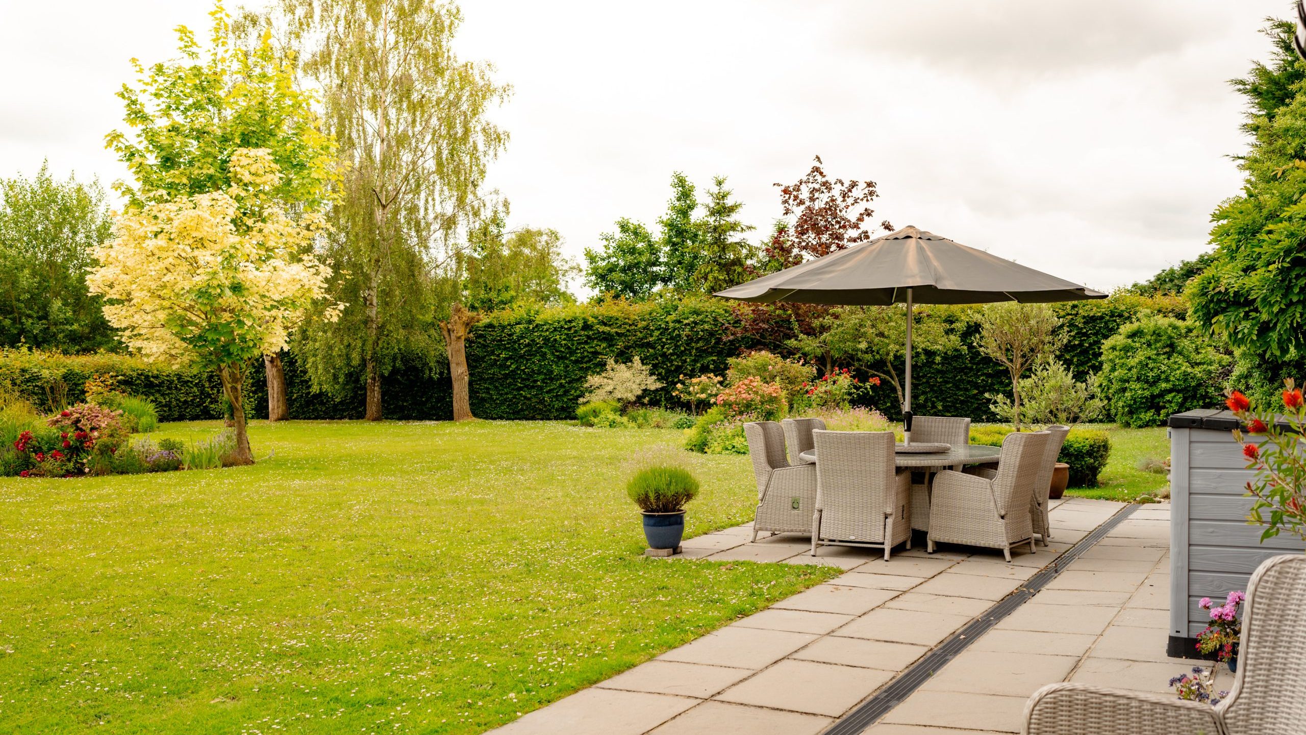 A well-maintained backyard featuring a patio with a round table, six cushioned chairs, and a large umbrella. The patio is bordered by lush greenery, various trees, and colorful bushes. The expansive lawn stretches out with scattered flower beds.