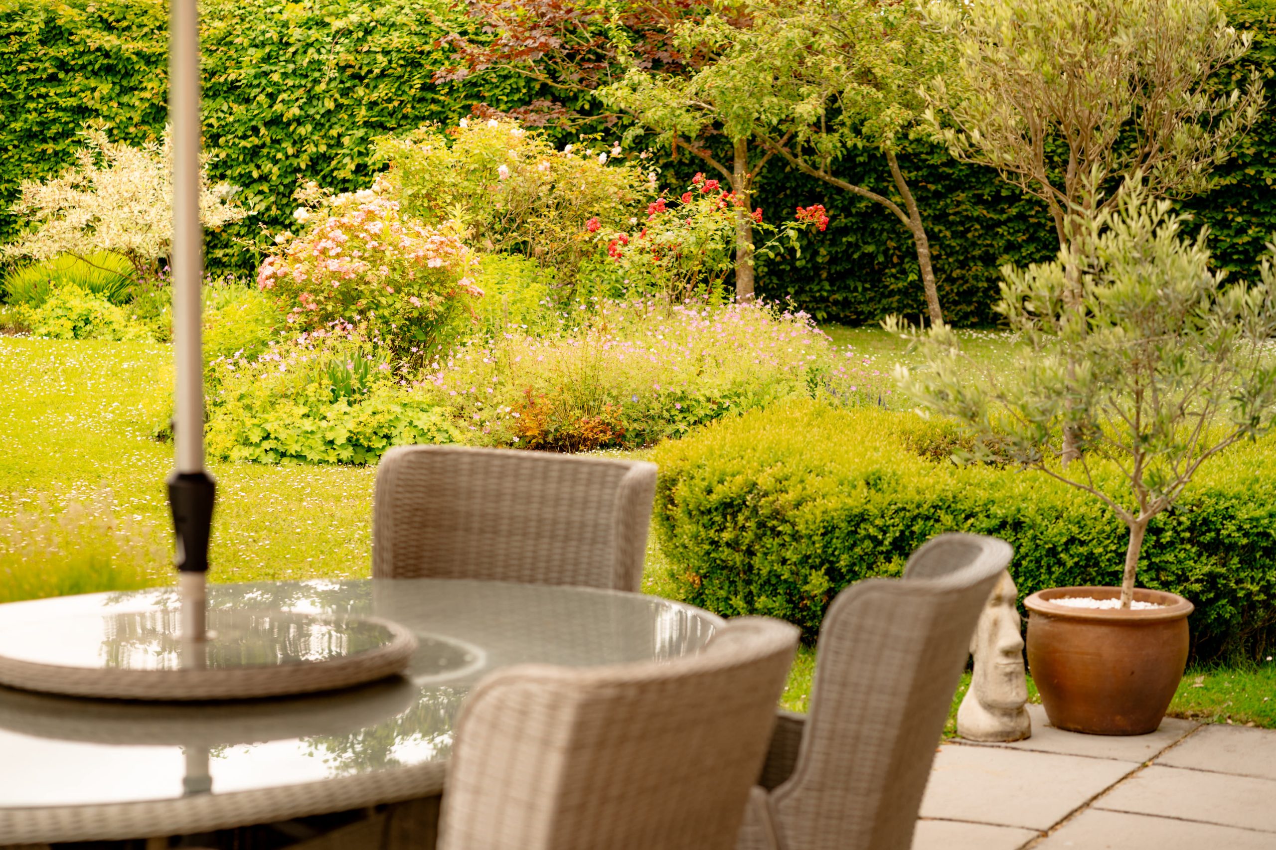 A backyard garden features a round glass-top table with four wicker chairs on a patio with slate tiles. The lush garden has various plants, shrubs, and blooming flowers surrounded by a green hedge. A small potted tree adds to the serene atmosphere.